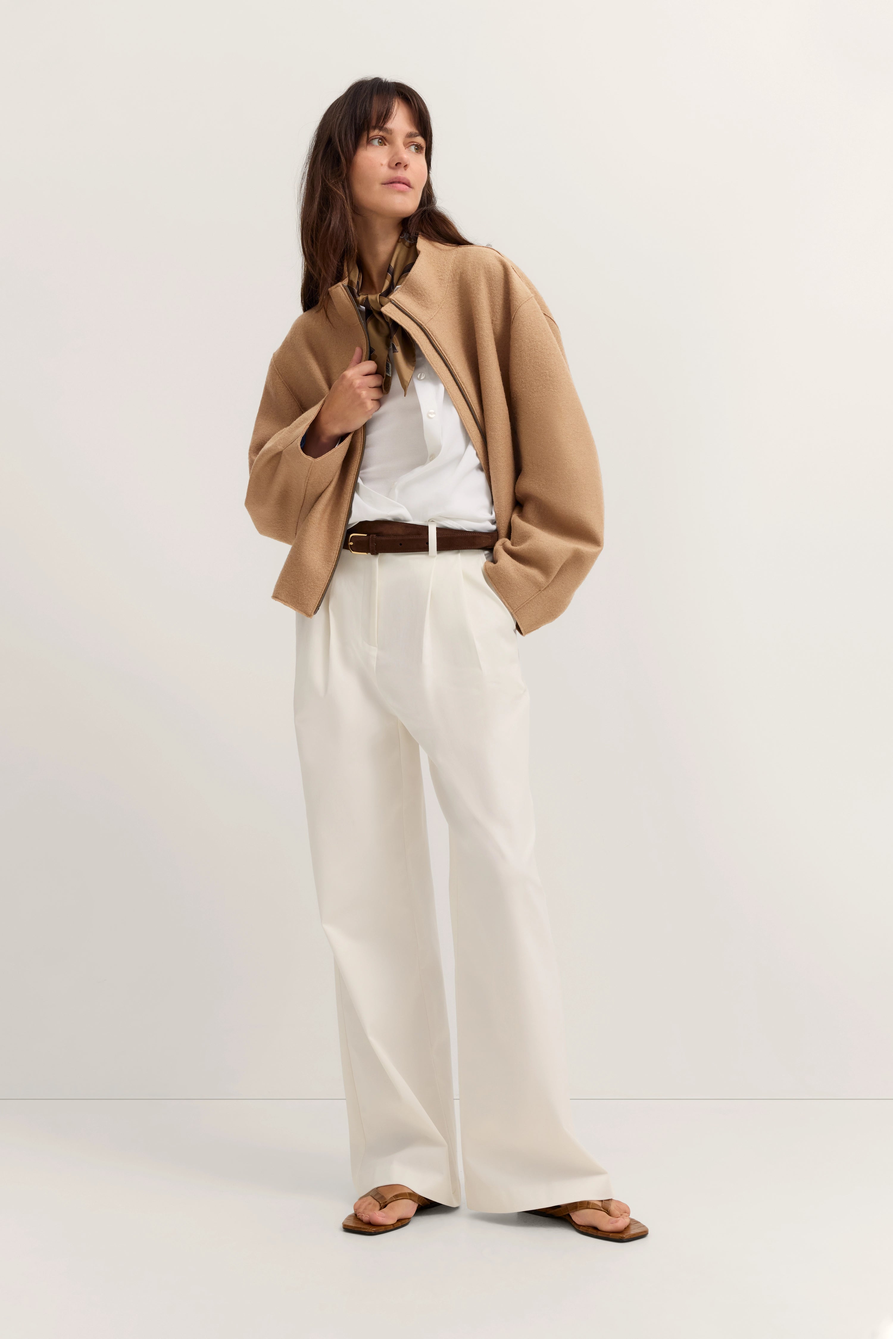 Model trägt beiges Cape, weiße Bluse, weite weiße Hose, braunen Gürtel. Lässige Pose.