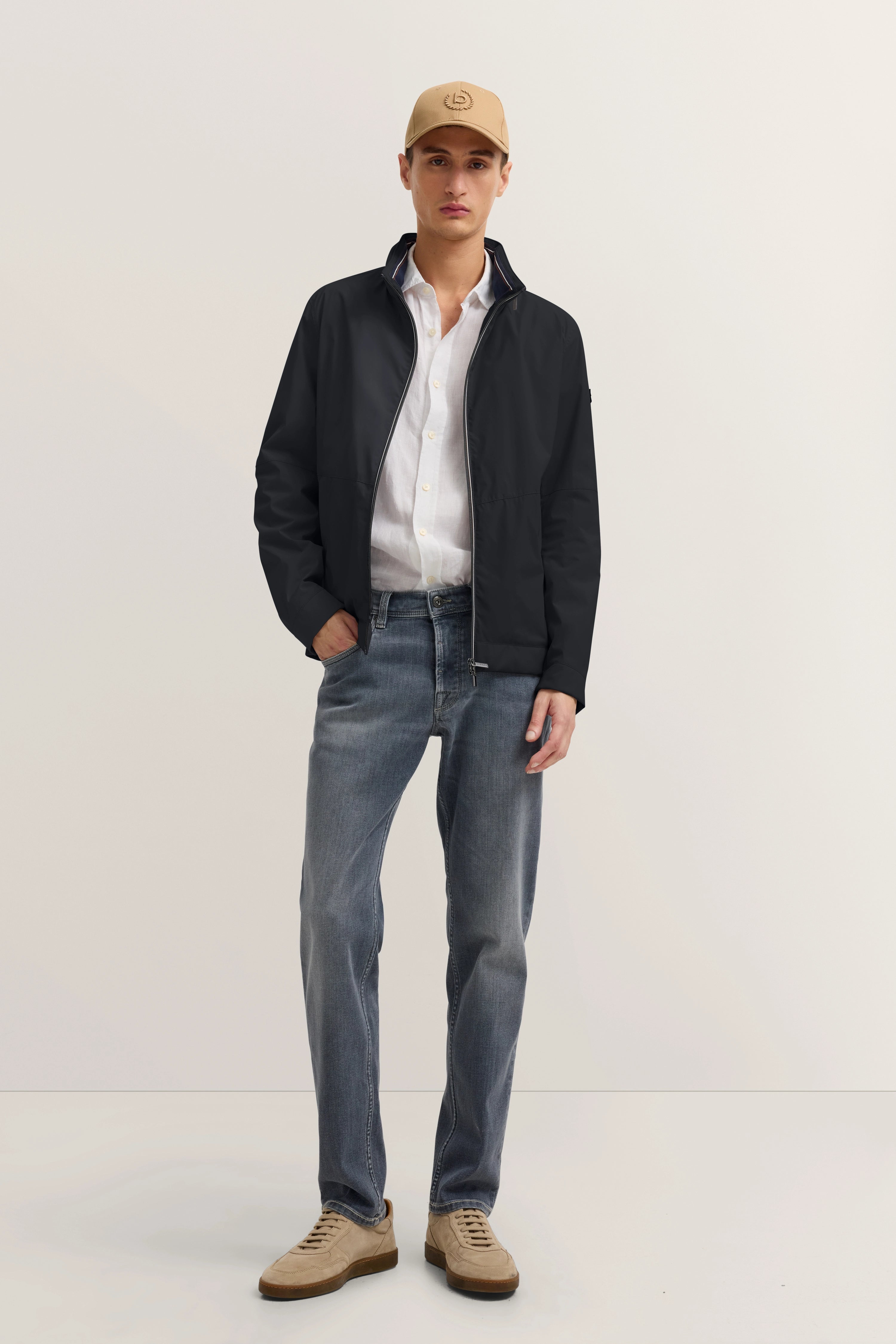 bugatti Model in schwarzer Jacke, weißem Hemd, blauer Jeans, beige Mütze und Schuhen, lässige Pose.