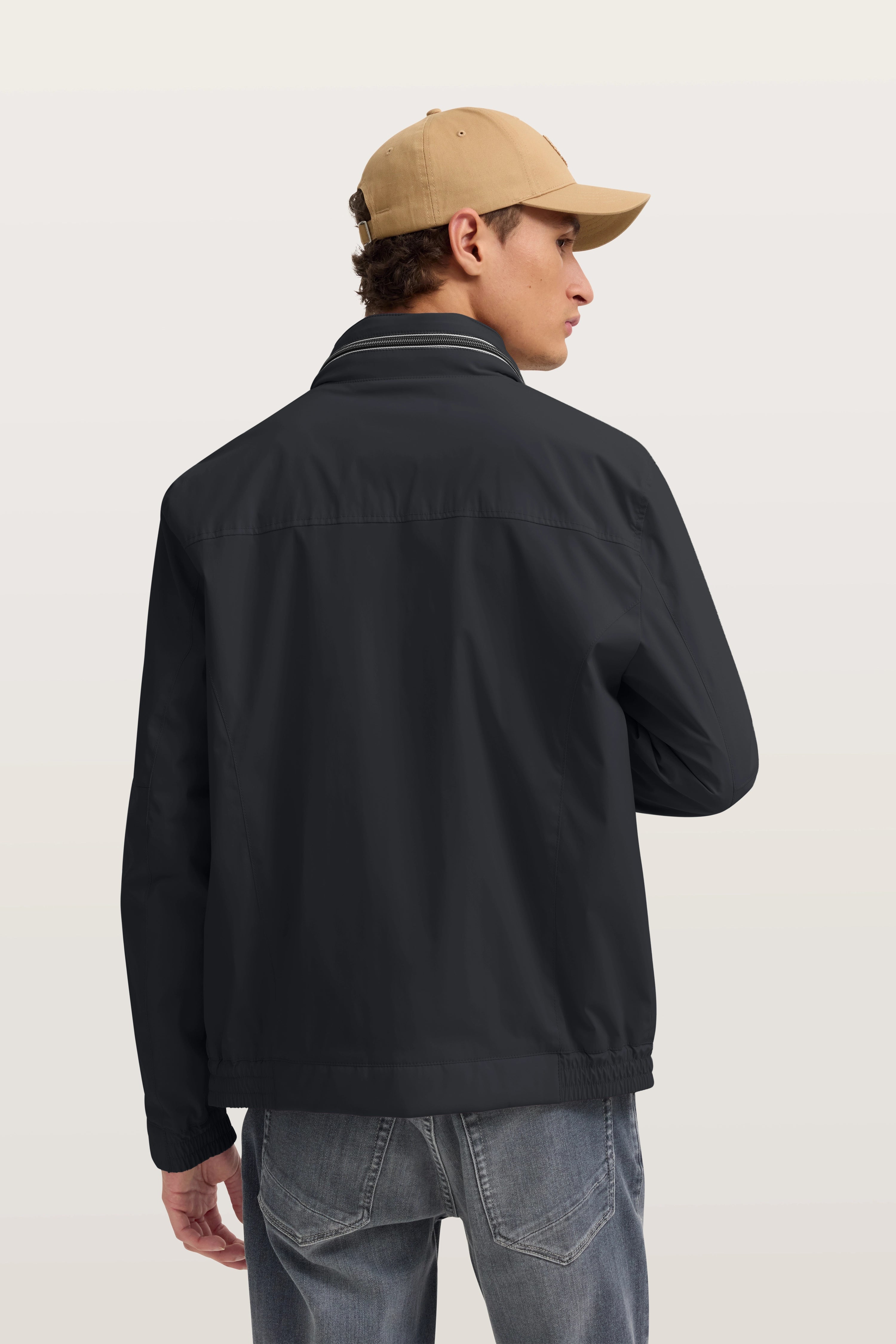 Model in schwarzer bugatti Jacke, beige Kappe, Jeans, Rückansicht, entspannte Pose.