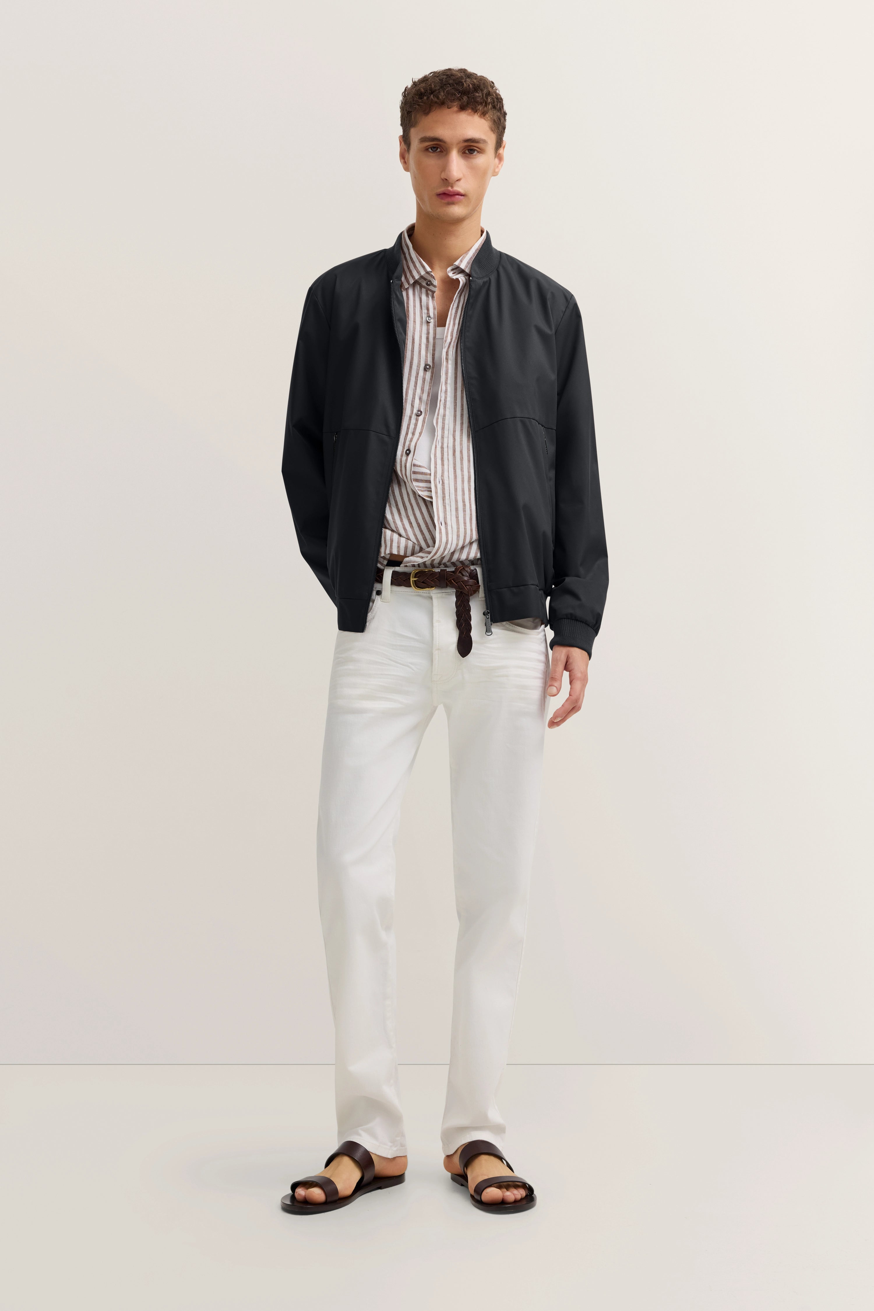 "bugatti Model in schwarzer Jacke, gestreiftem Hemd, weißen Hosen. Lässig stehend mit entspannter Pose."
