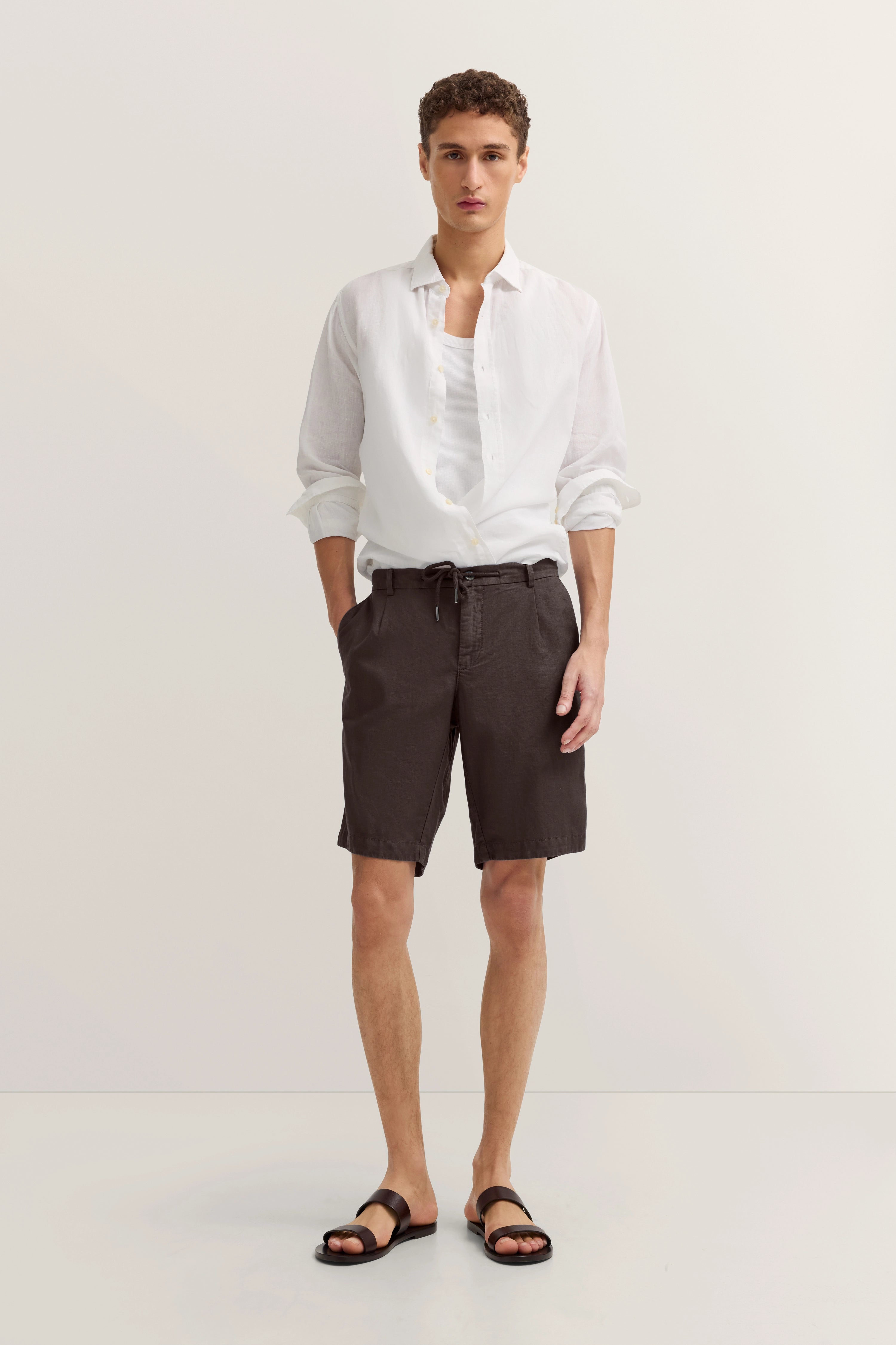 Weißes Hemd, braune Shorts, Model steht entspannt, Arme in Taschen, bugatti Stil und Geschmack.
