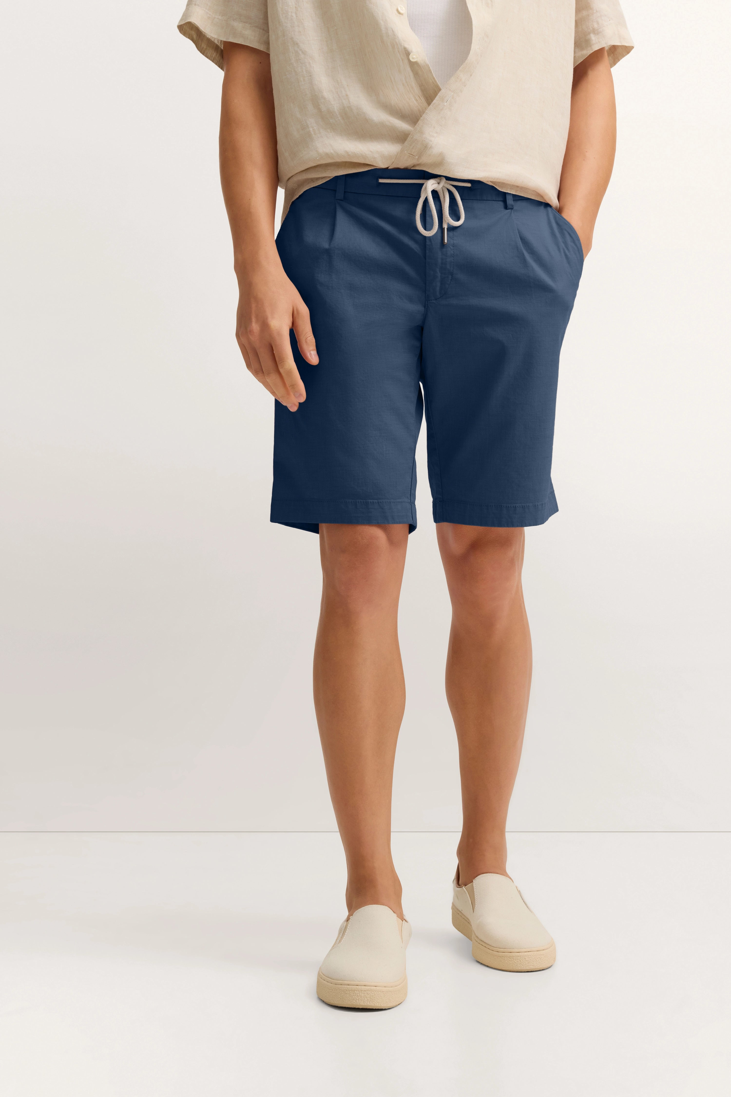 Beige Hemd mit blauen Shorts und beigen Schuhen, legeres Modelbild, Hände in Taschen.
