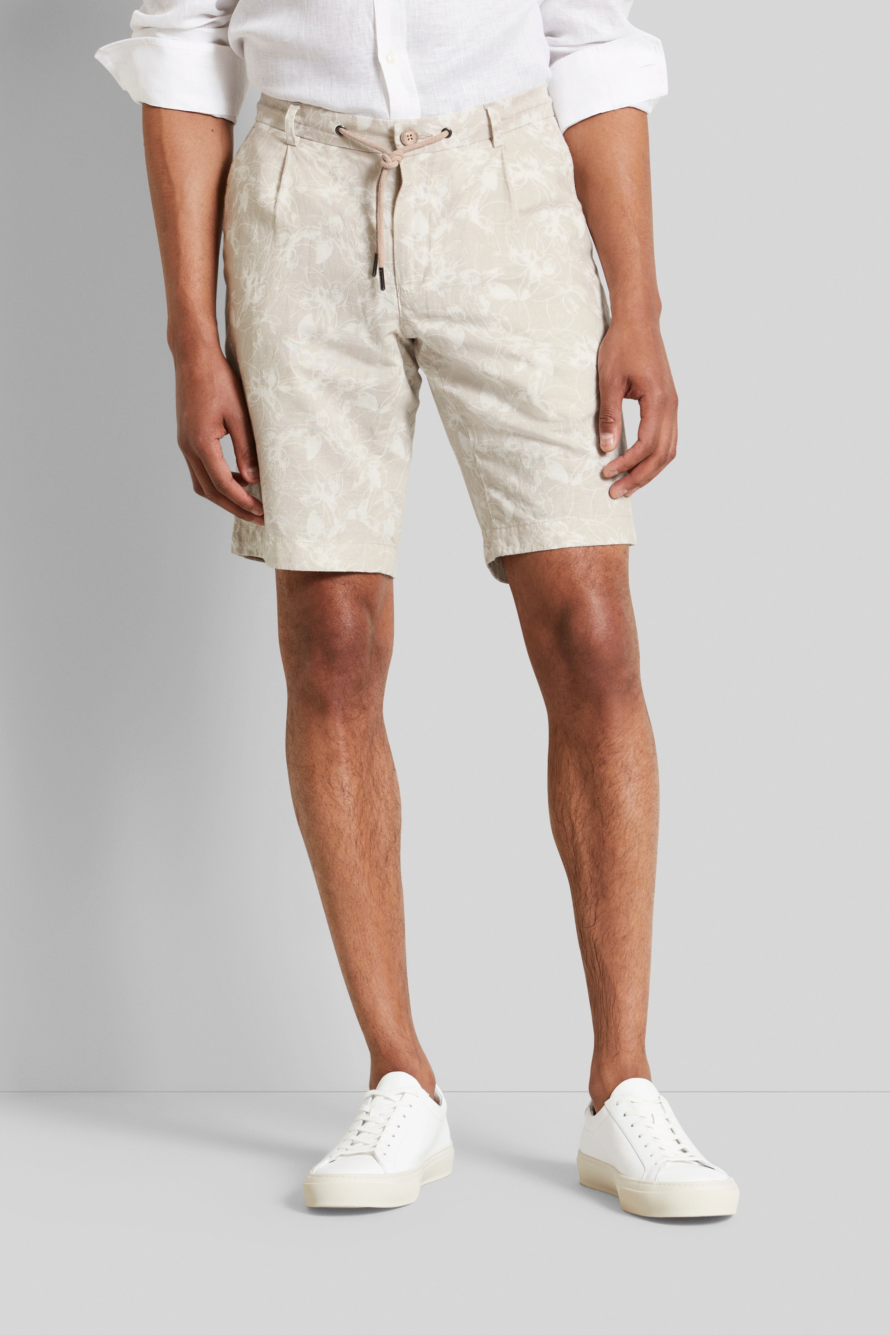 Beige gemusterte Shorts mit weißem Hemd und Sneakers, legeres Modelbild, entspannte Pose.
