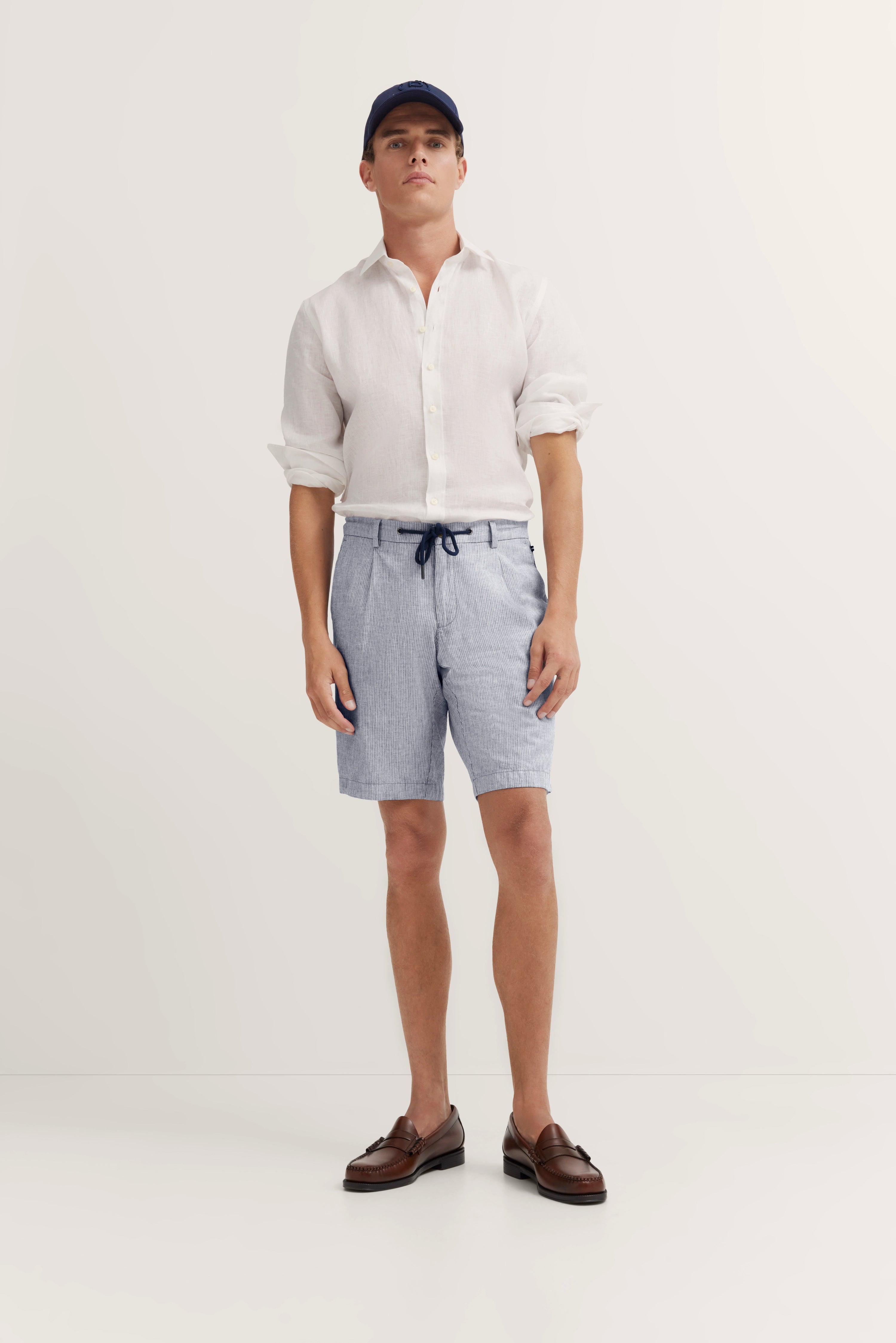 Model in weißem Hemd, blauen Shorts und braunen Schuhen, entspanntes Posing.