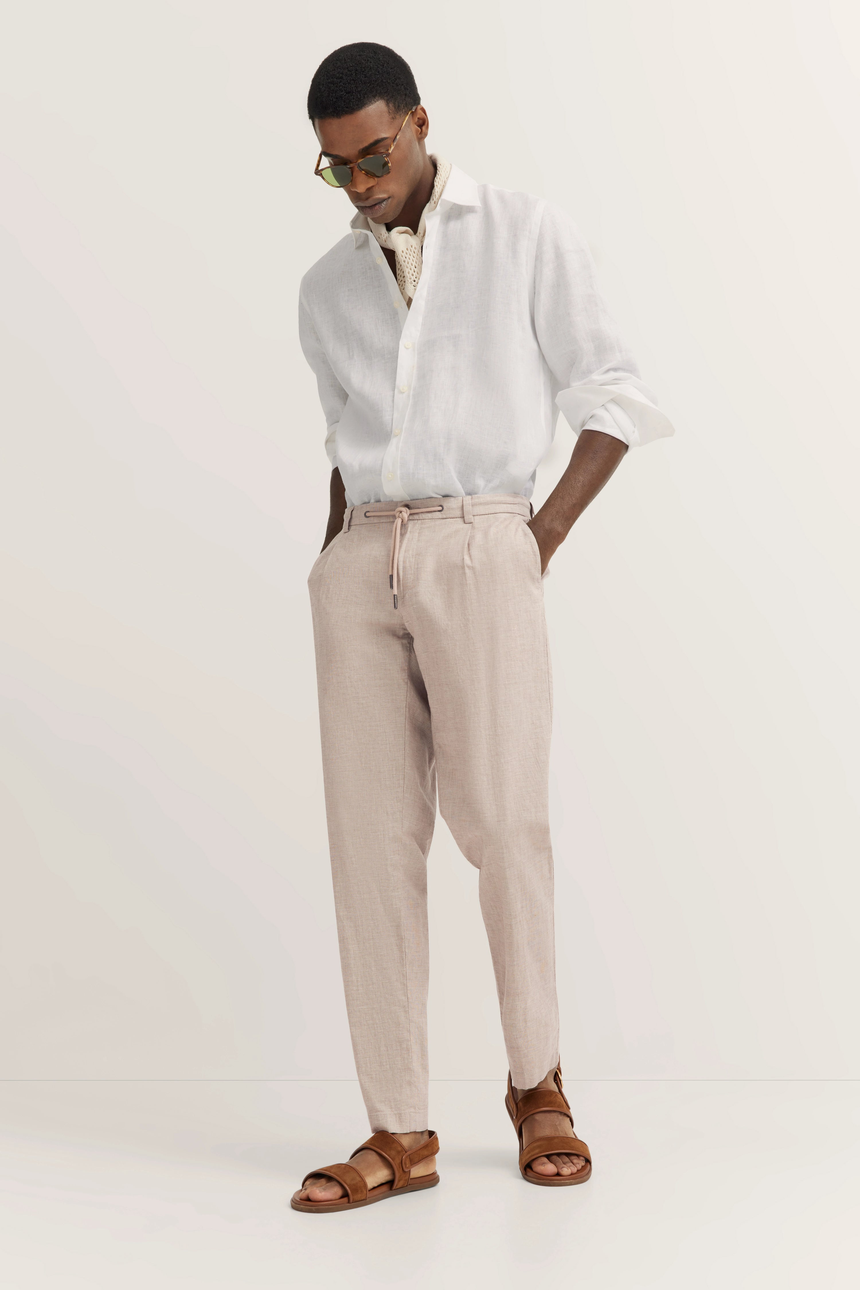 Witte linnen blouse, beige broek, bruine sandalen van bugatti. Model poseert ontspannen, handen in zakken.