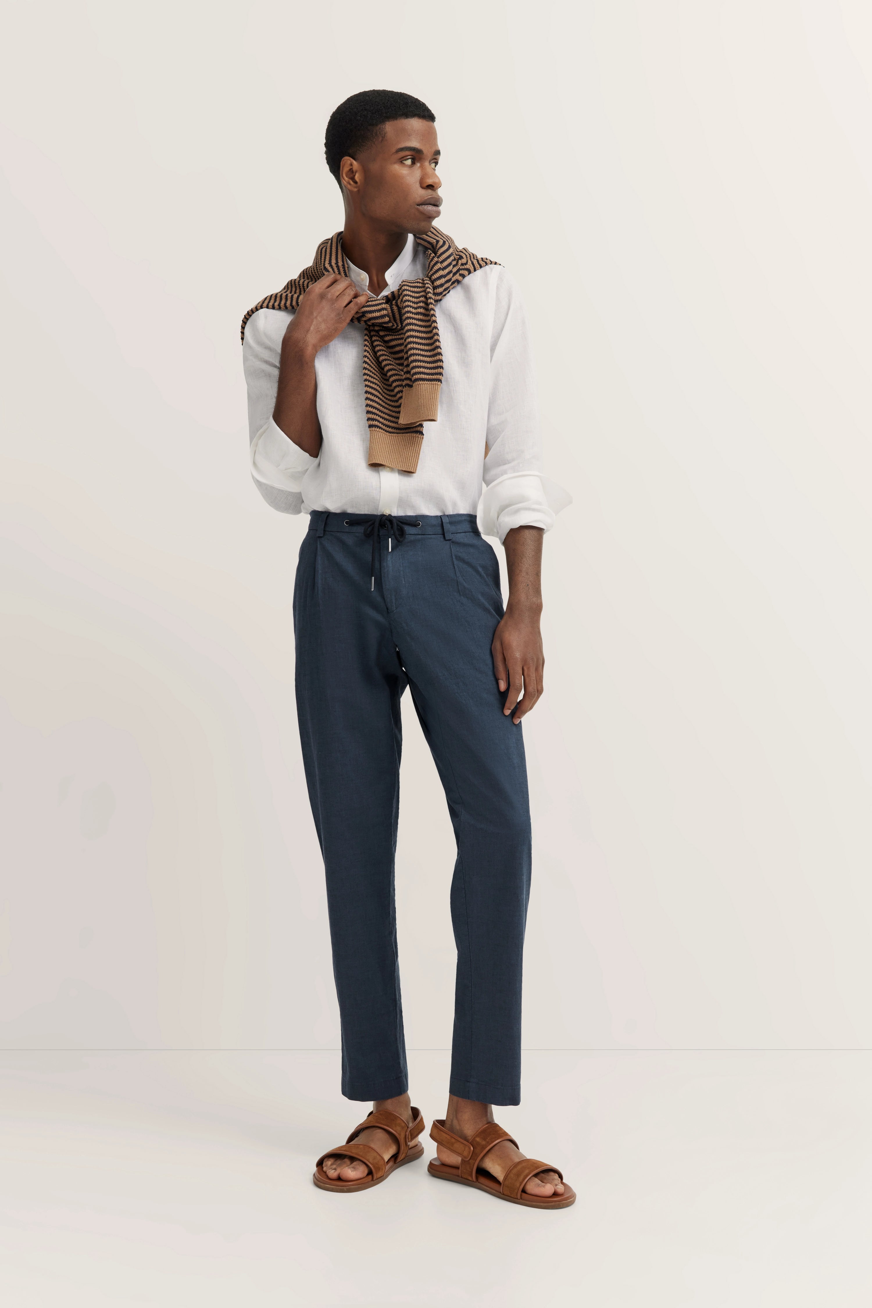 Model in wit overhemd, blauwe broek, gestreepte sjaal, bruine sandalen. Ontspannen pose. bugatti-stijl.