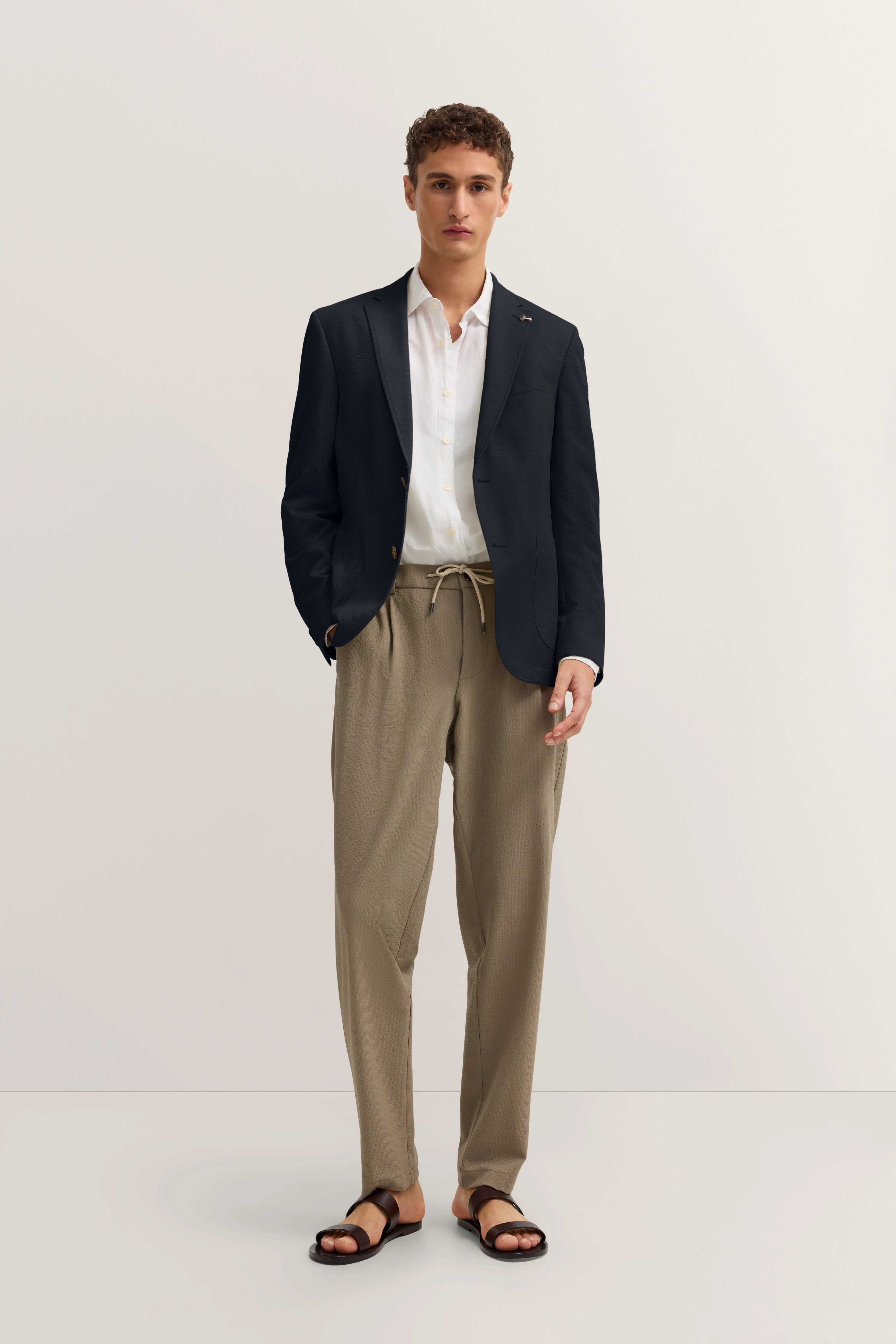 bugatti Modelbild: Dunkler Blazer, weißes Hemd, beige Hose, schwarze Sandalen, entspanntes Posing.