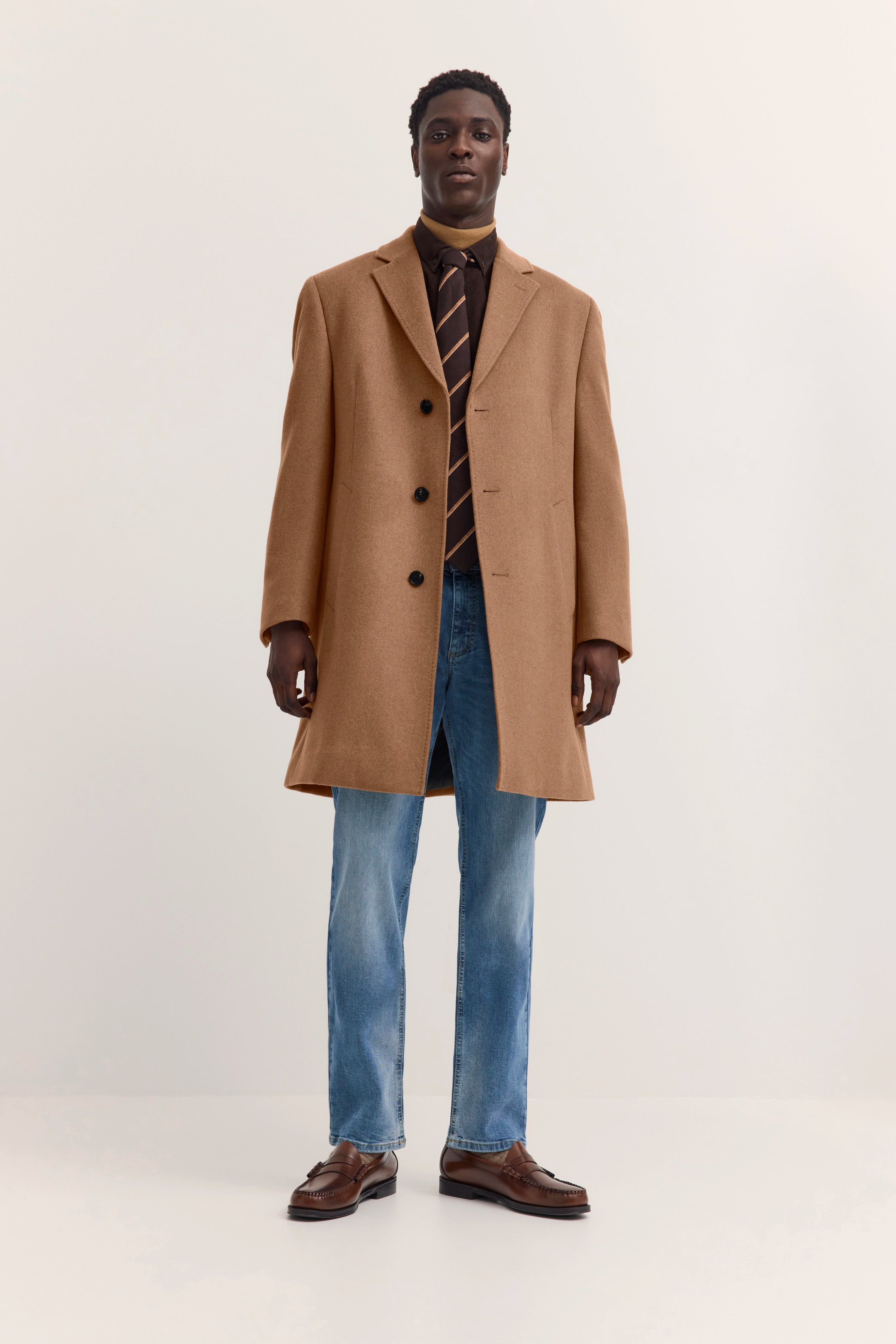 Modelbild mit beigem Mantel, braunem Hemd, gestreifter Krawatte, blauer Jeans und braunen Schuhen, frontal.