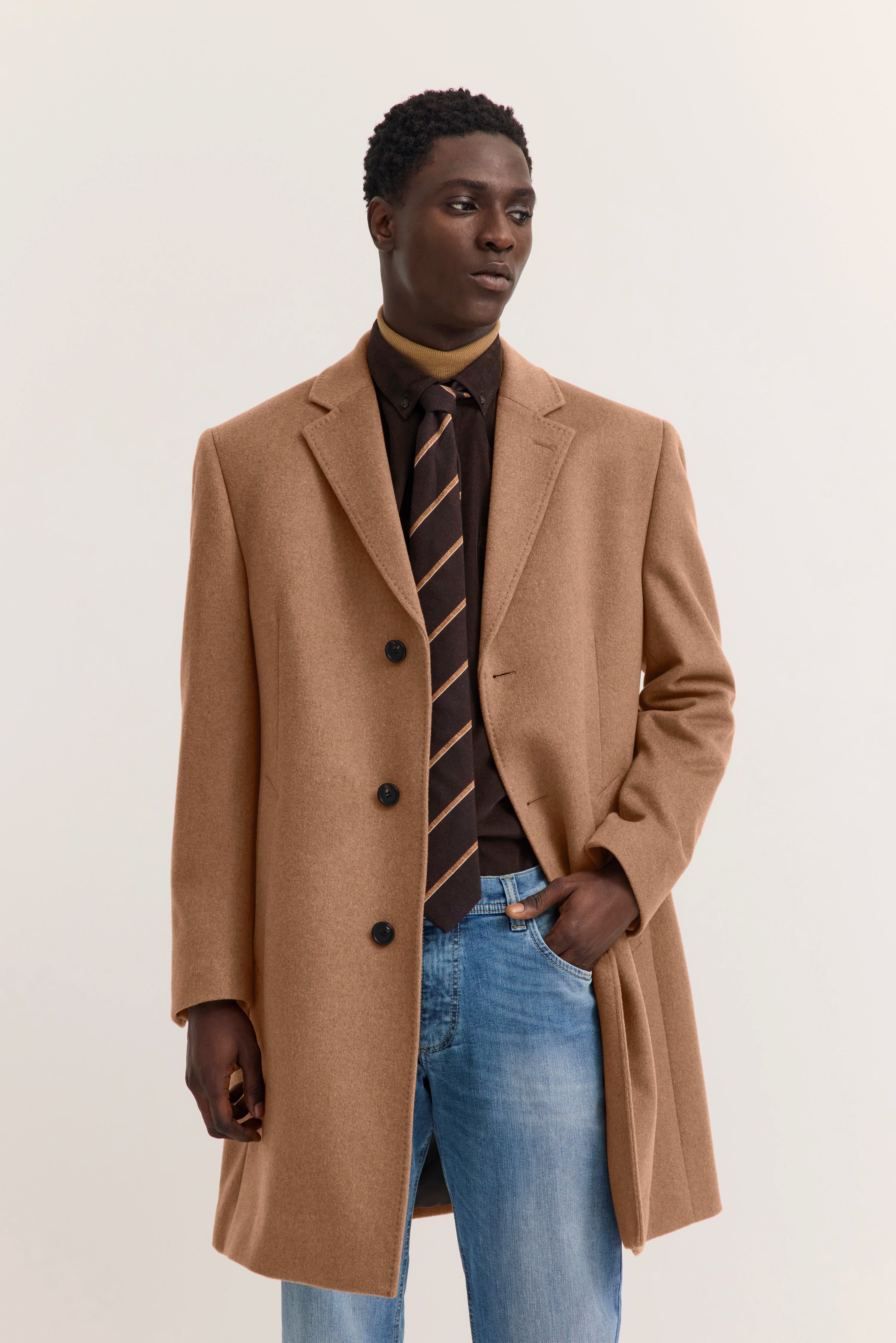 Model in camel coat, brown shirt, striped tie, blue jeans. Lässig mit Hand in Tasche. Bugatti Stil.