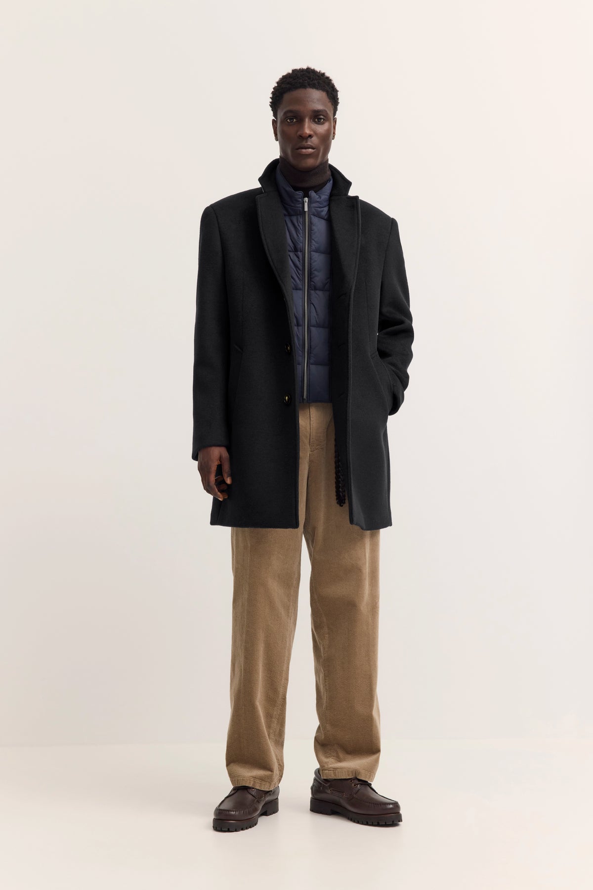 Model im schwarzen Mantel, blauer Steppjacke, beige Cordhose