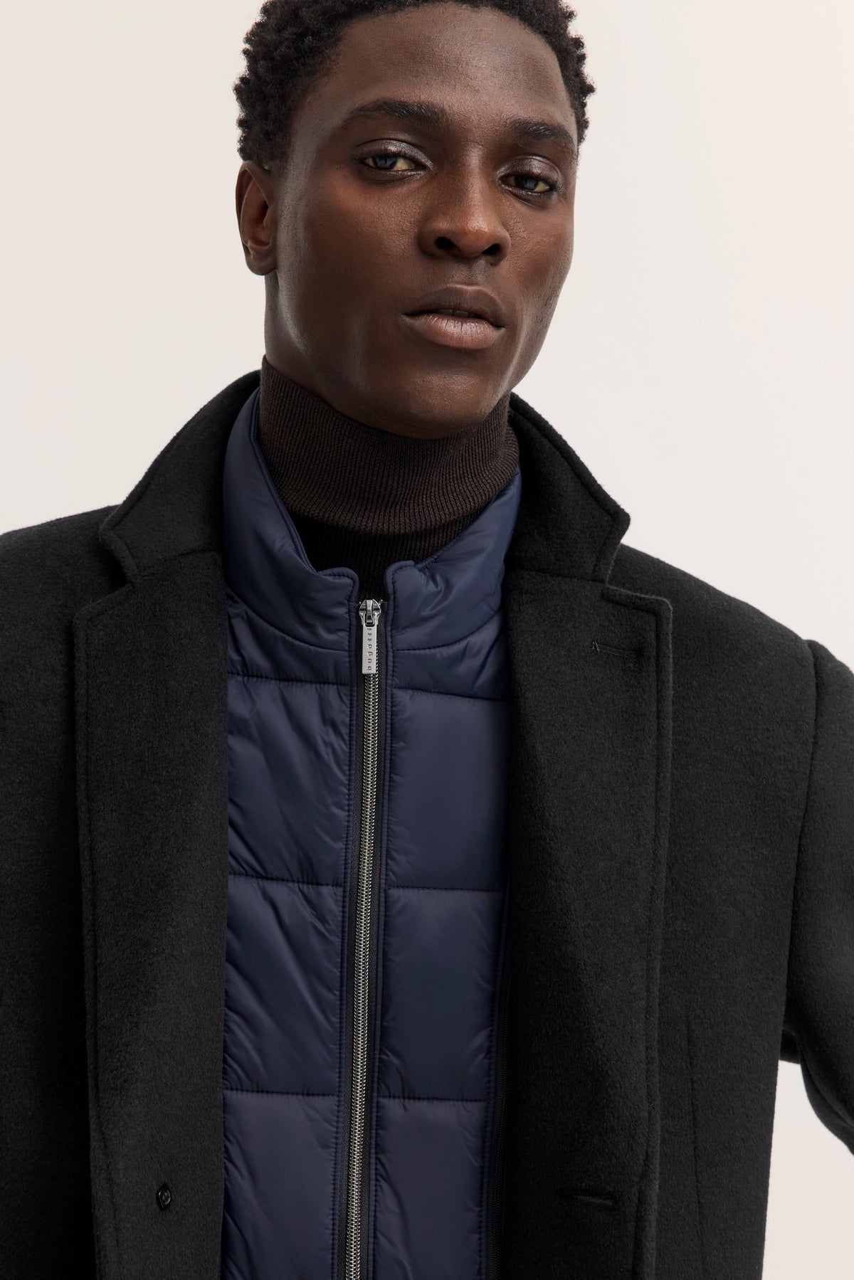 Schwarzer Mantel, blauer Steppjacke, schwarzer Rollkragen, Modelbild, entspannte Pose.