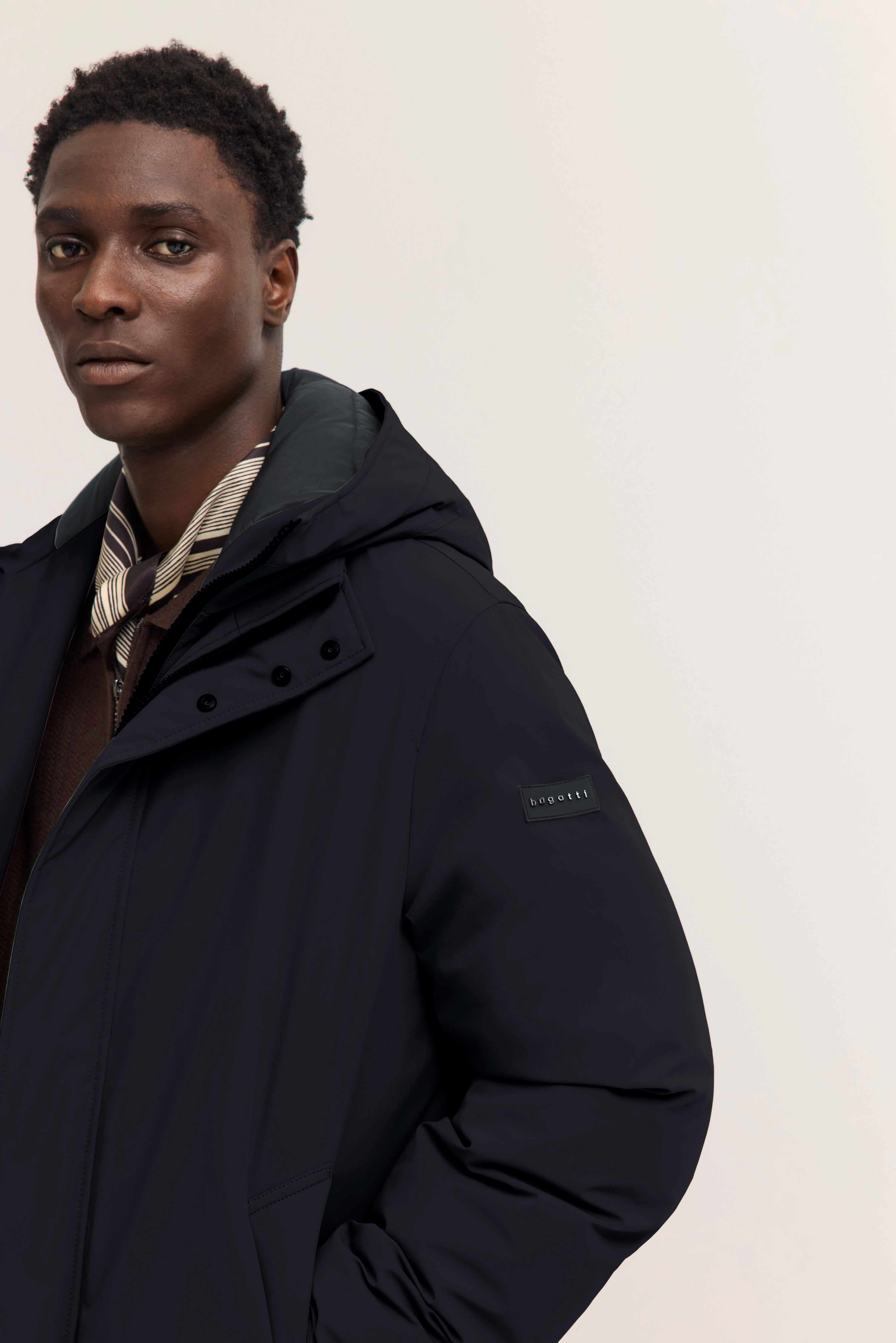 Bugatti Model in schwarzer Jacke, braune Strickjacke, gestreifte Krawatte, entspanntes Posing.