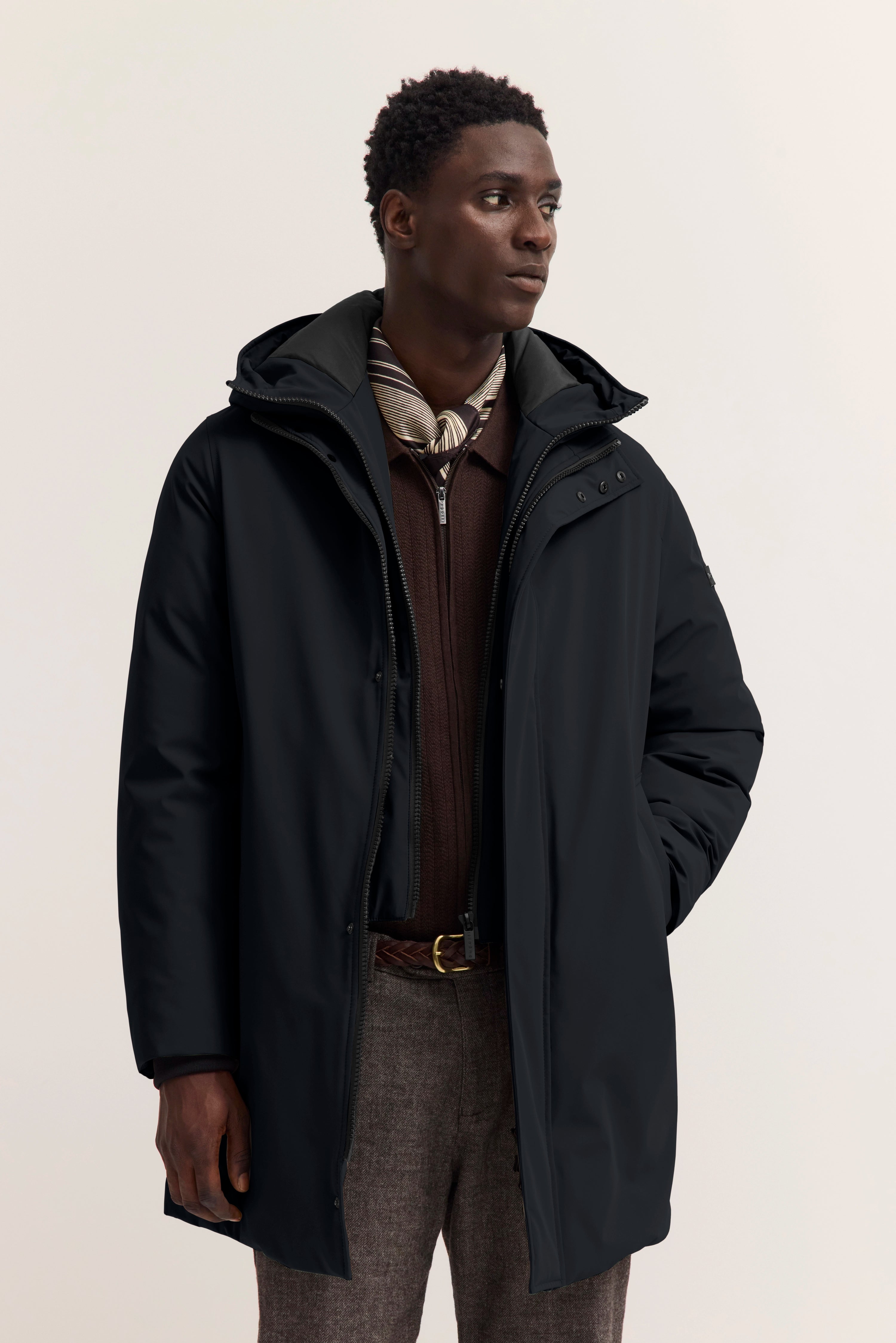 bugatti Modelbild: Schwarzer Mantel, brauner Pullover, gemusterter Schal. Stehende, elegante Pose.