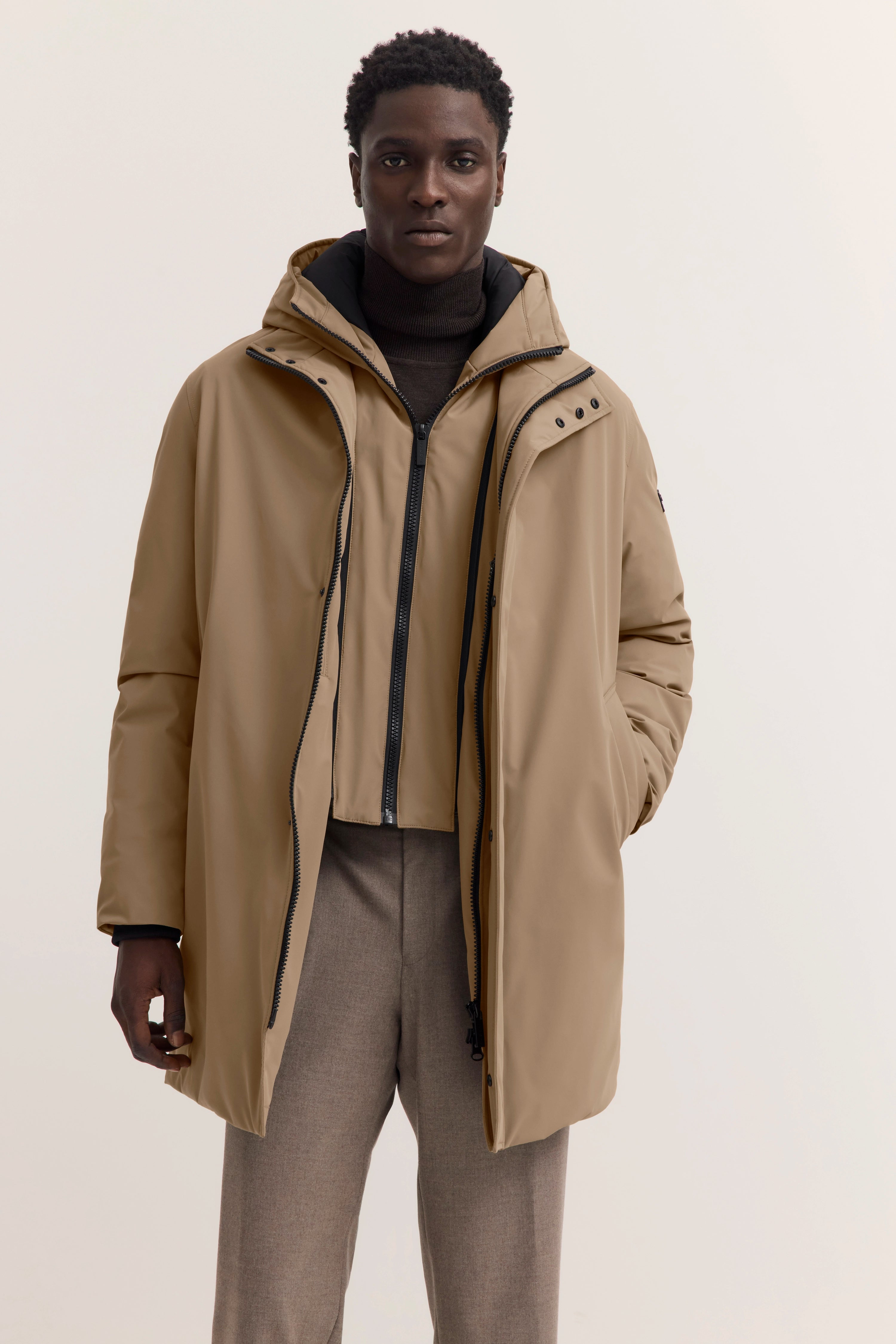 Beige bugatti Mantel, schwarzer Rollkragen, beige Hose, Model frontal, Hände in Taschen.