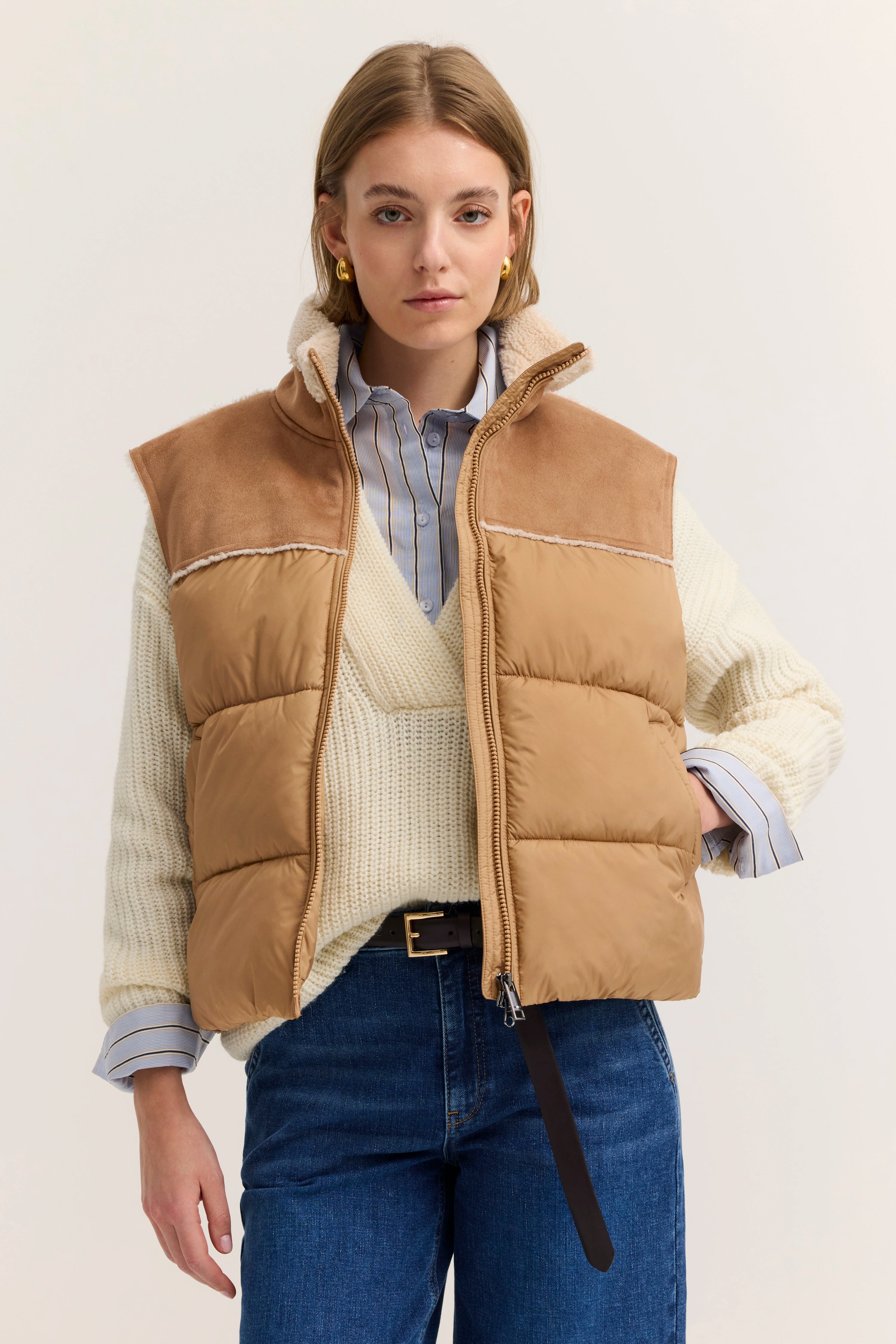 Model trägt beige Weste über gestreiftem Hemd und cremefarbenem Pullover, Jeans, entspannte Pose.