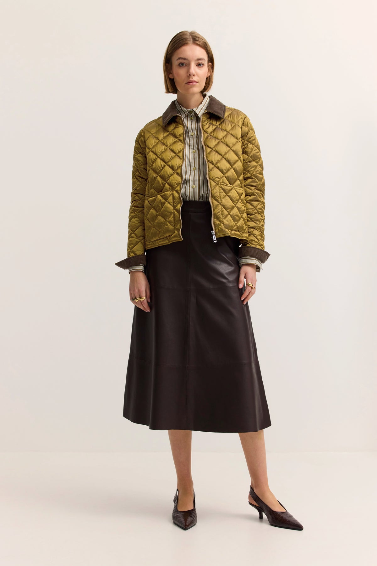 Model trägt gesteppte, olivgrüne Jacke, gestreifte Bluse, braunen Lederrock, spitze Schuhe. Entspanntes Posing.