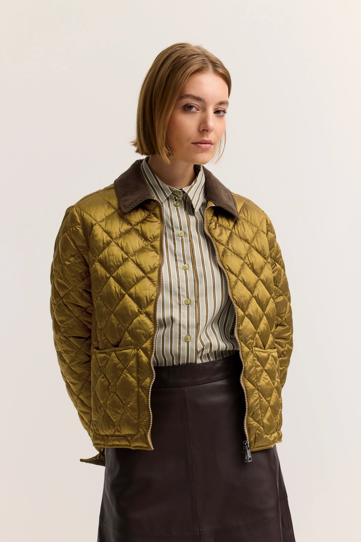Model in gestepptem, goldenen bugatti Blouson, gestreiftes Hemd, brauner Rock, entspannte Pose.