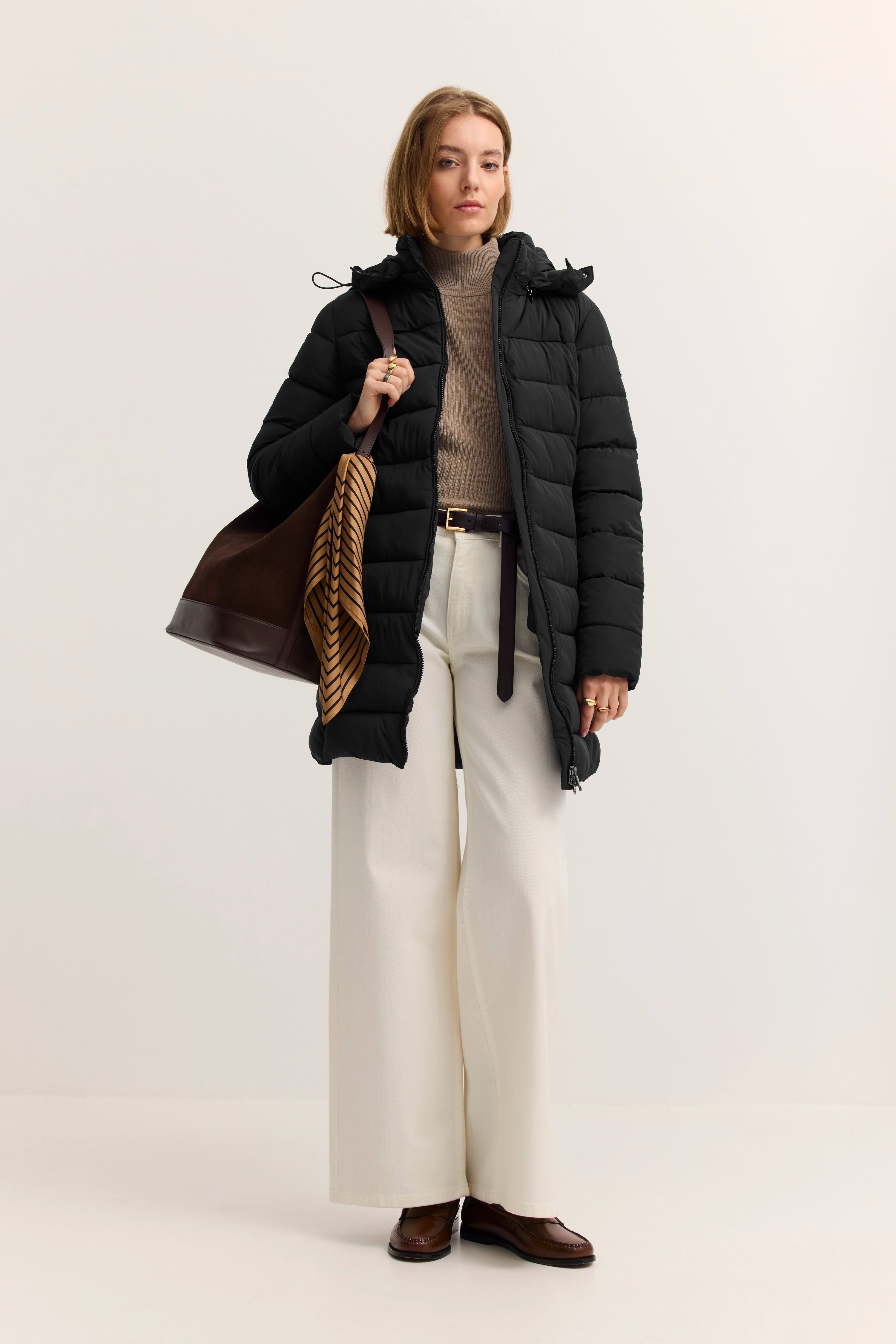 Model in schwarzem bugatti Daunenmantel, beiger Rollkragenpullover, weiße Hose, braune Schuhe, lässige Pose.