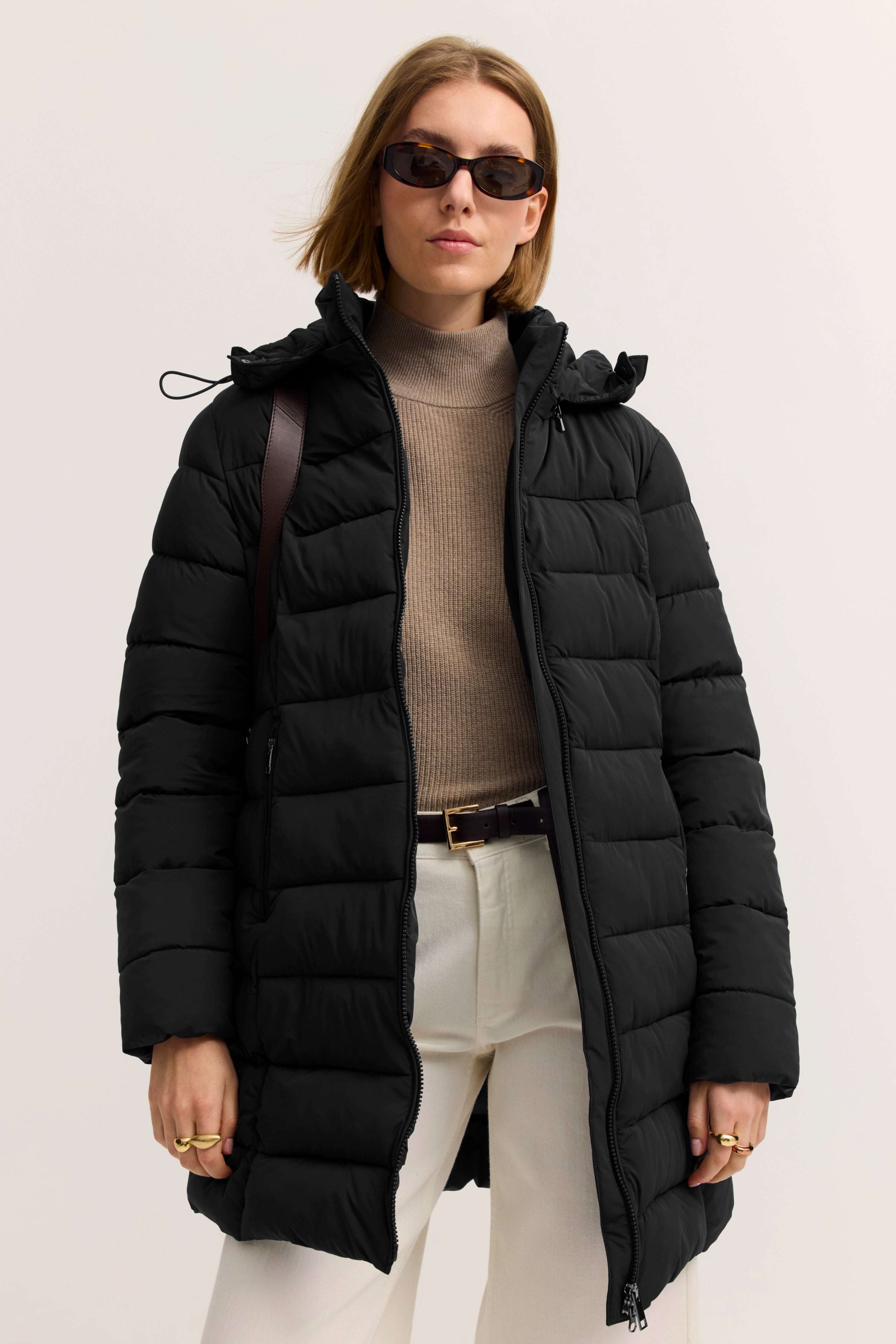 Model im schwarzen Daunenmantel, beigem Rollkragenpullover, weißer Hose. Lässig-elegant mit Sonnenbrille.