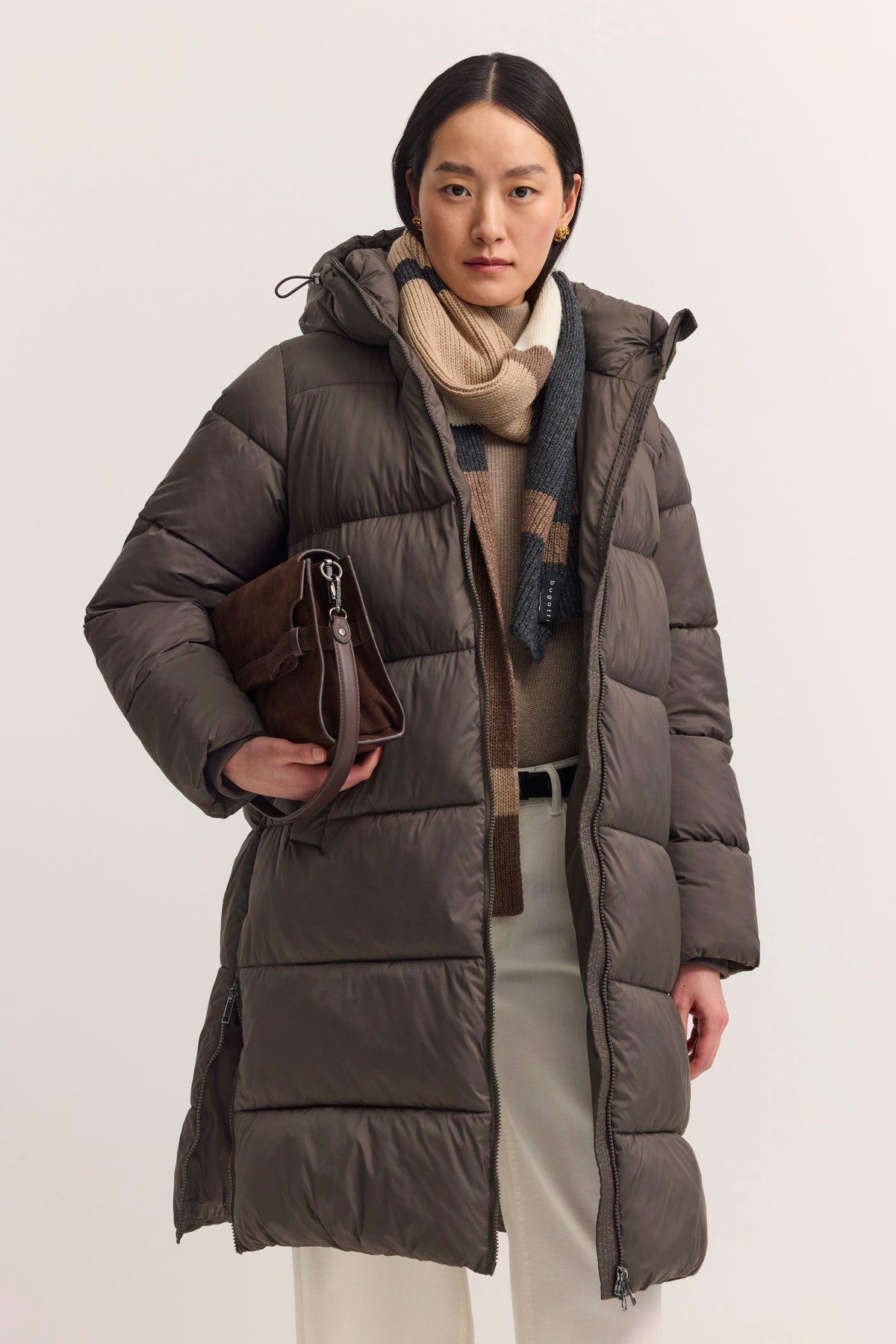 Model in braunem bugatti Steppmantel, beiger Pullover, gestreifter Schal, weiße Hose, braune Tasche.