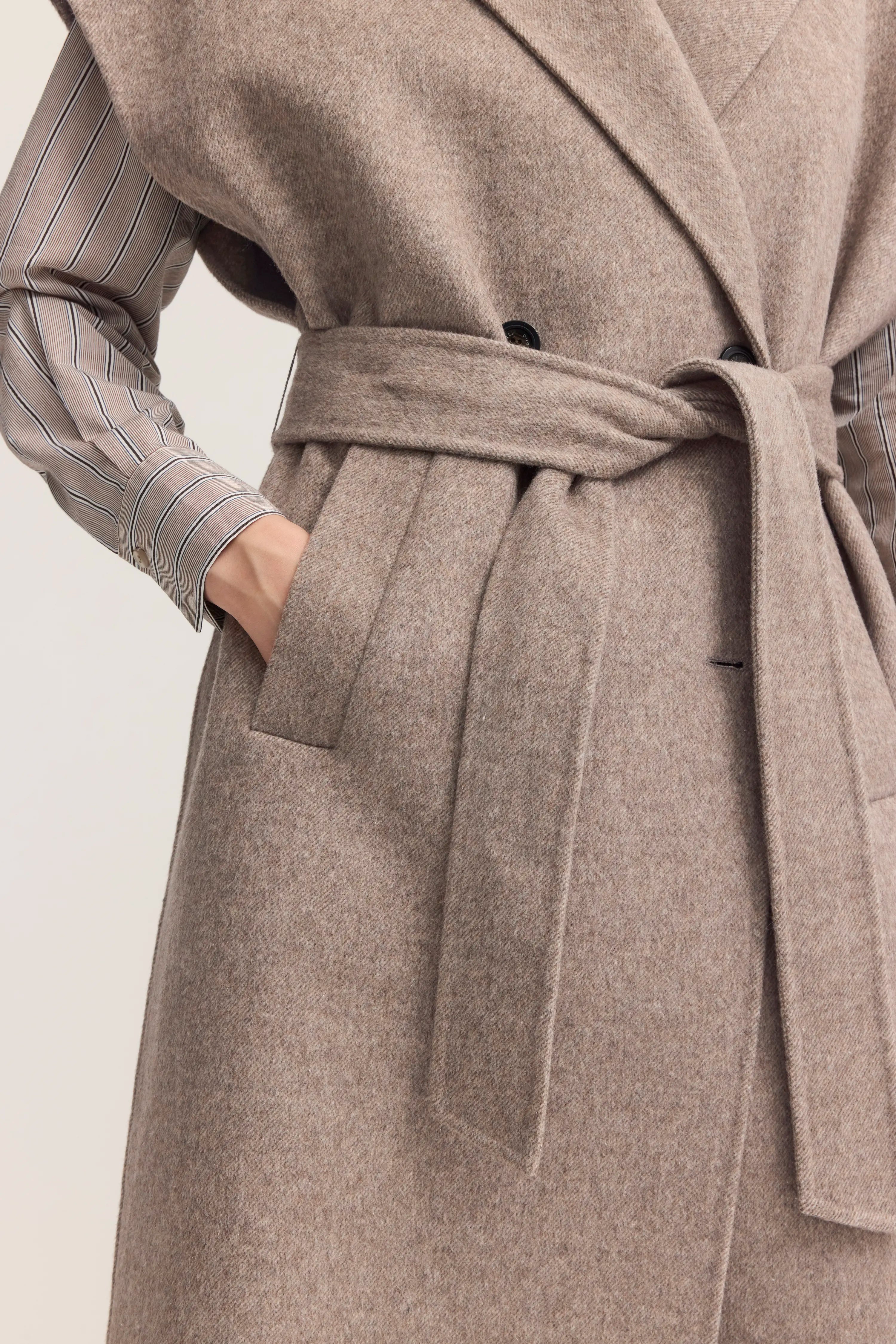 Strukturierter Mantel in Beige mit Gürtel, auf Büstenbild. Darunter gestreifte Bluse in Grau und Weiß.
