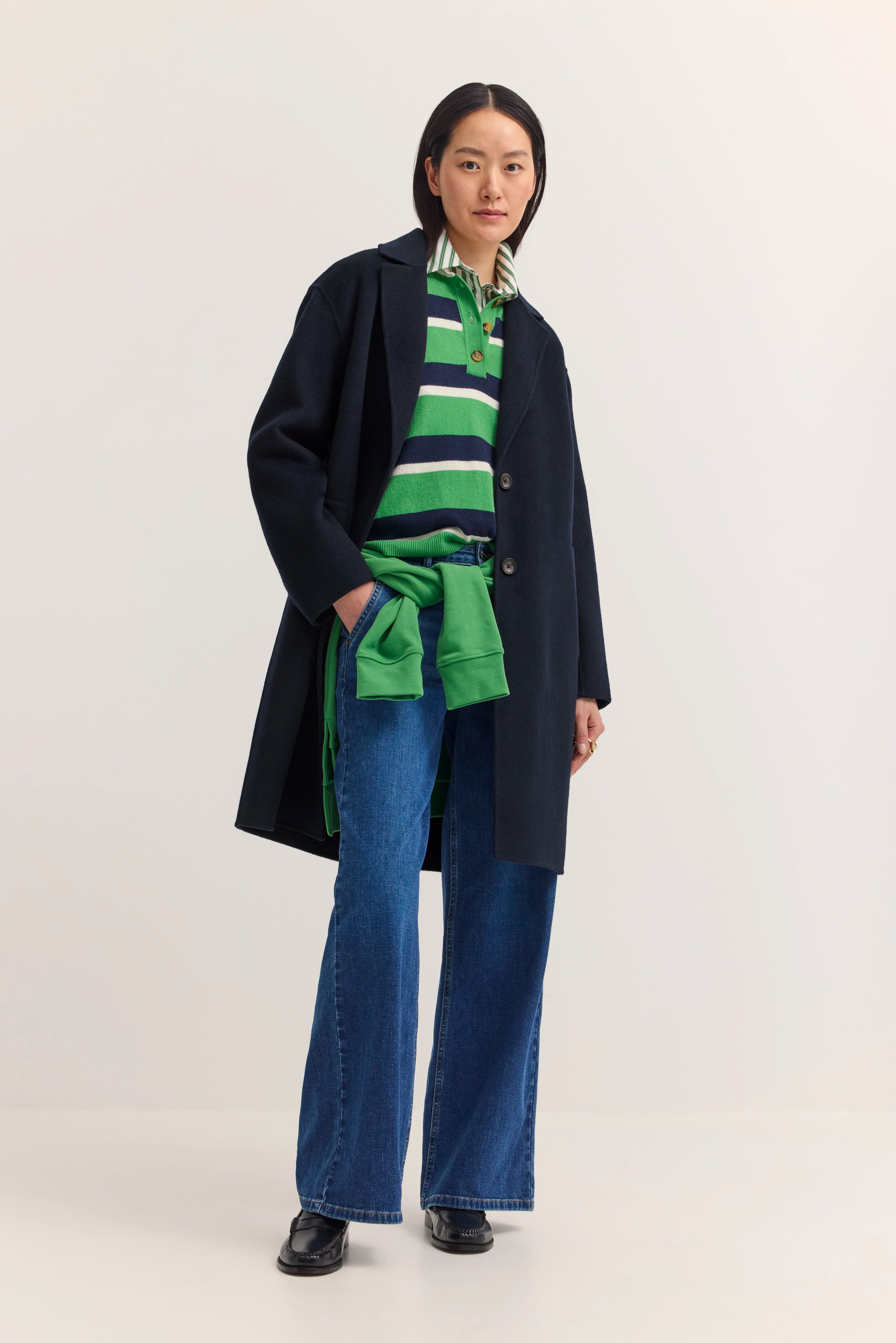 Model in grün gestreiftem Pullover, dunkelblauem Mantel, Jeans. Lässig mit Händen in den Taschen.