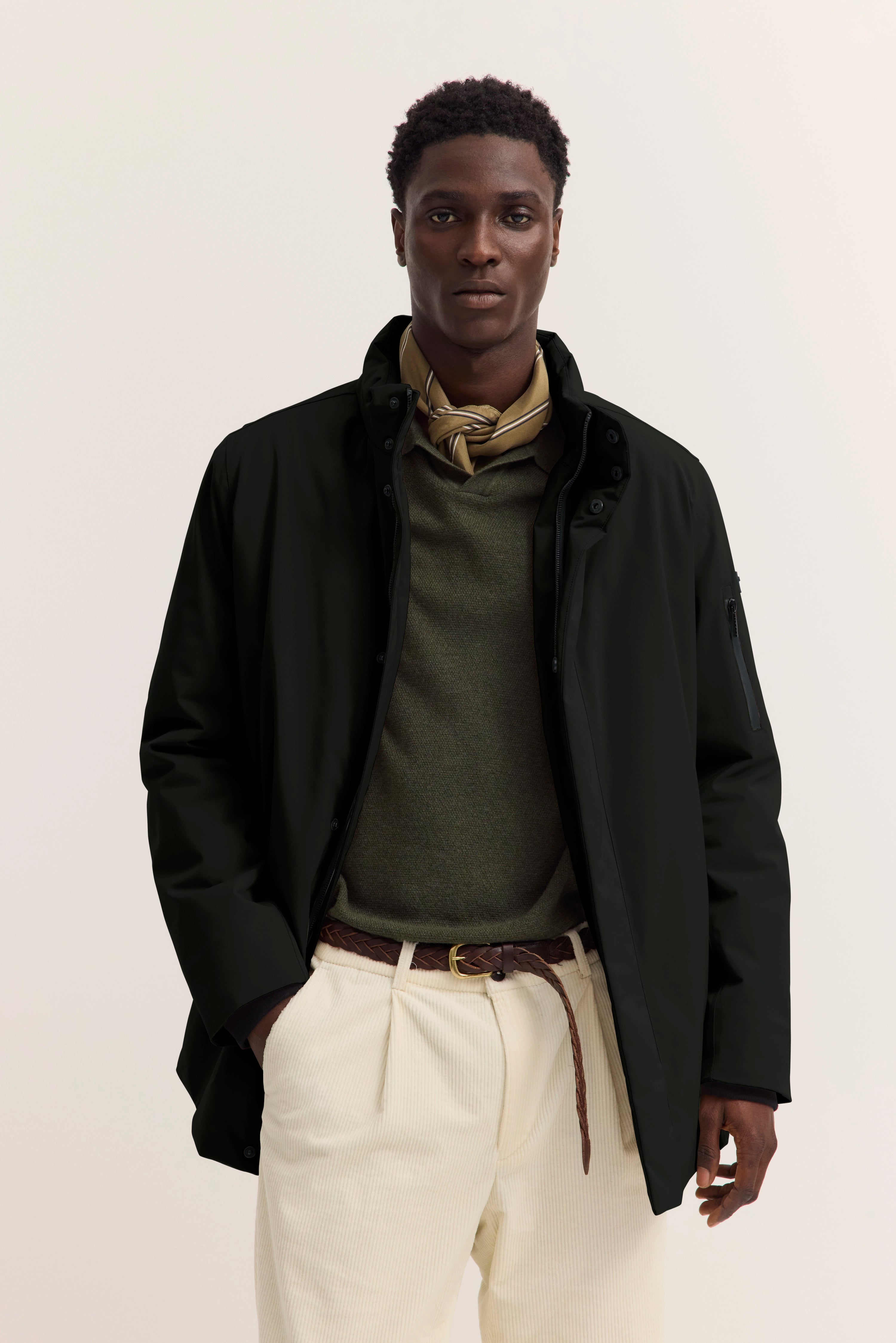 Model in schwarzer Jacke, olivgrünem Pullover, cremefarbenen Hosen, braunem Gürtel