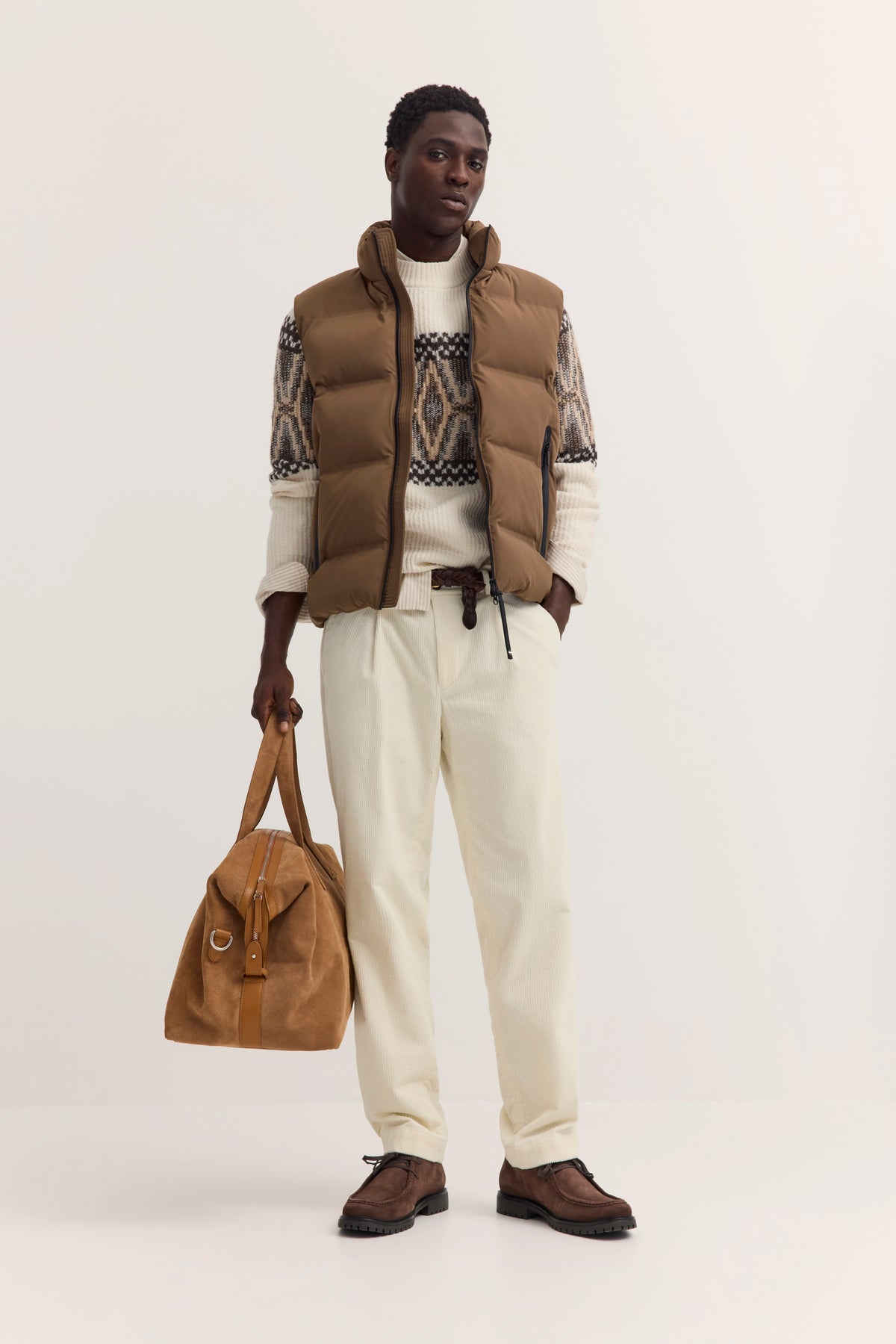 Braune Weste, gemusterter Pullover, beige Hose. Model steht mit Tasche.