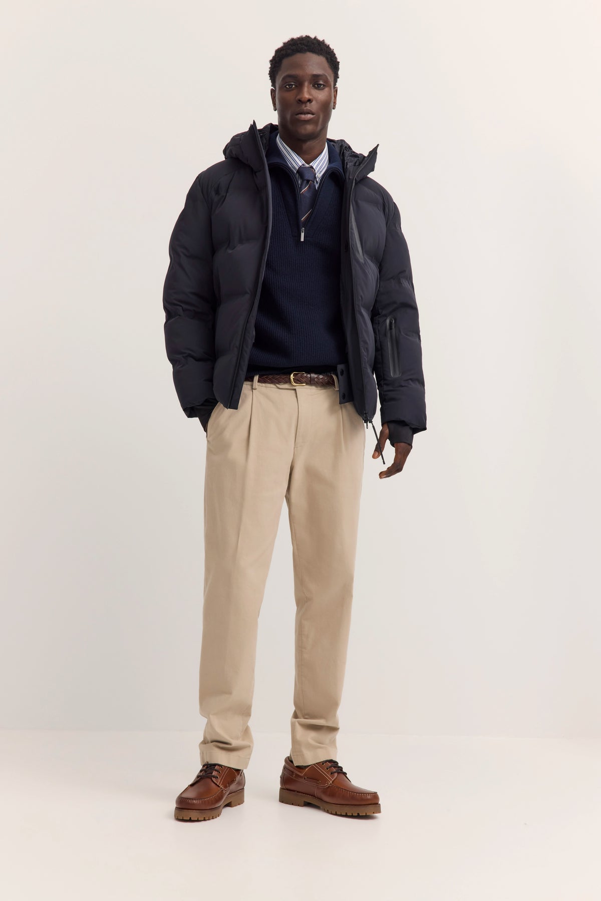 Model in schwarzer bugatti Daunenjacke, blauem Pullover, gestreiftem Hemd, beigefarbenen Chinos, braunen Schuhen.