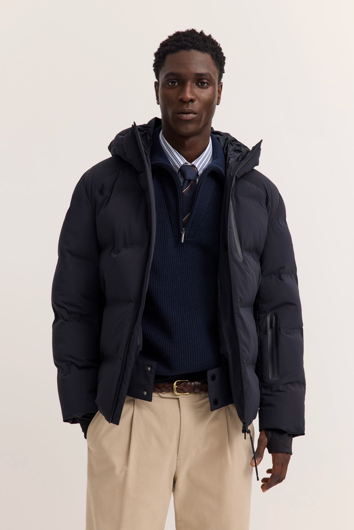 Model trägt blaue bugatti Daunenjacke, gestreiftes Hemd, marineblauen Pullover, beige Hose.