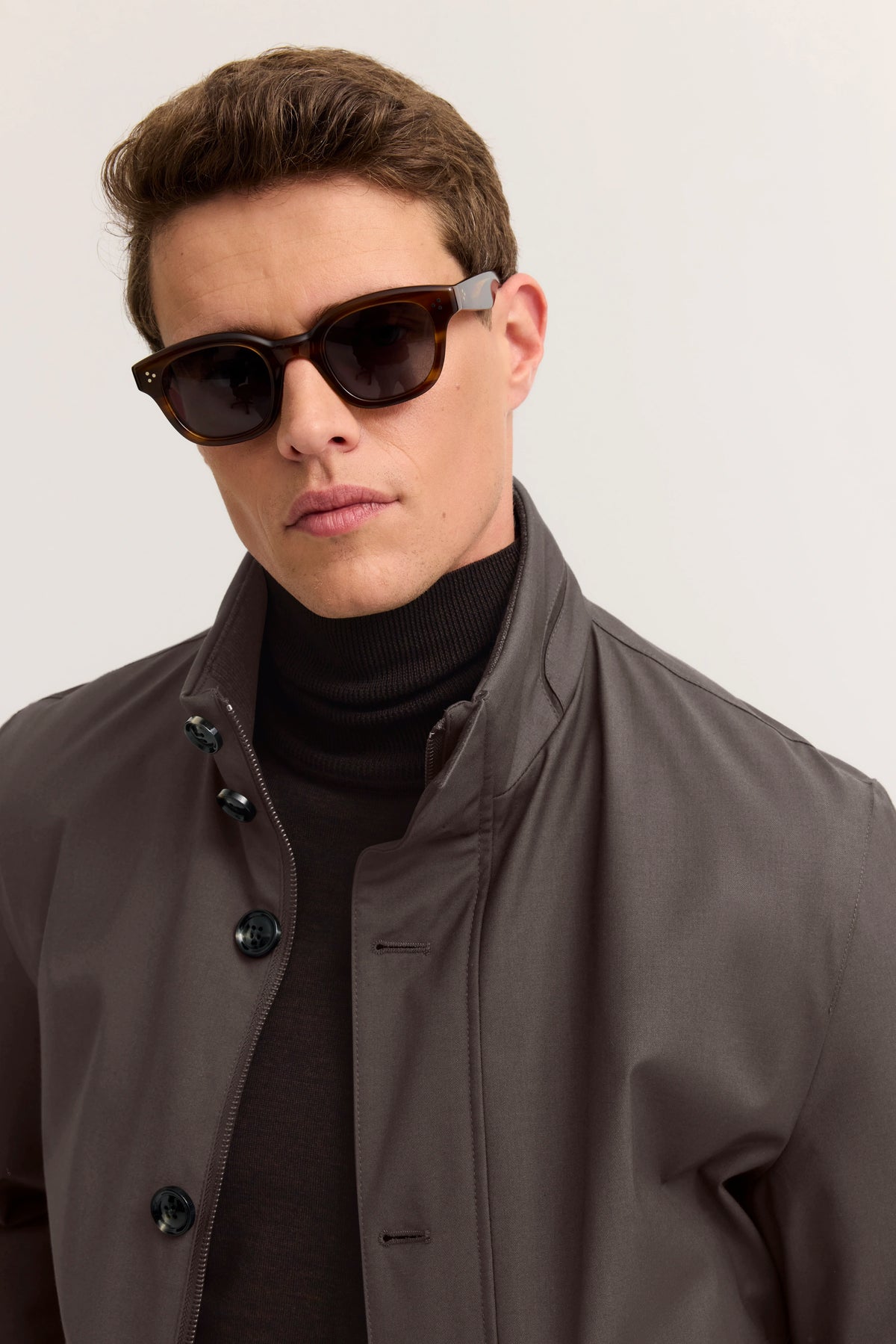 Model in grauem bugatti-Mantel und schwarzem Rollkragenpullover, trägt Sonnenbrille, Halbportrait.