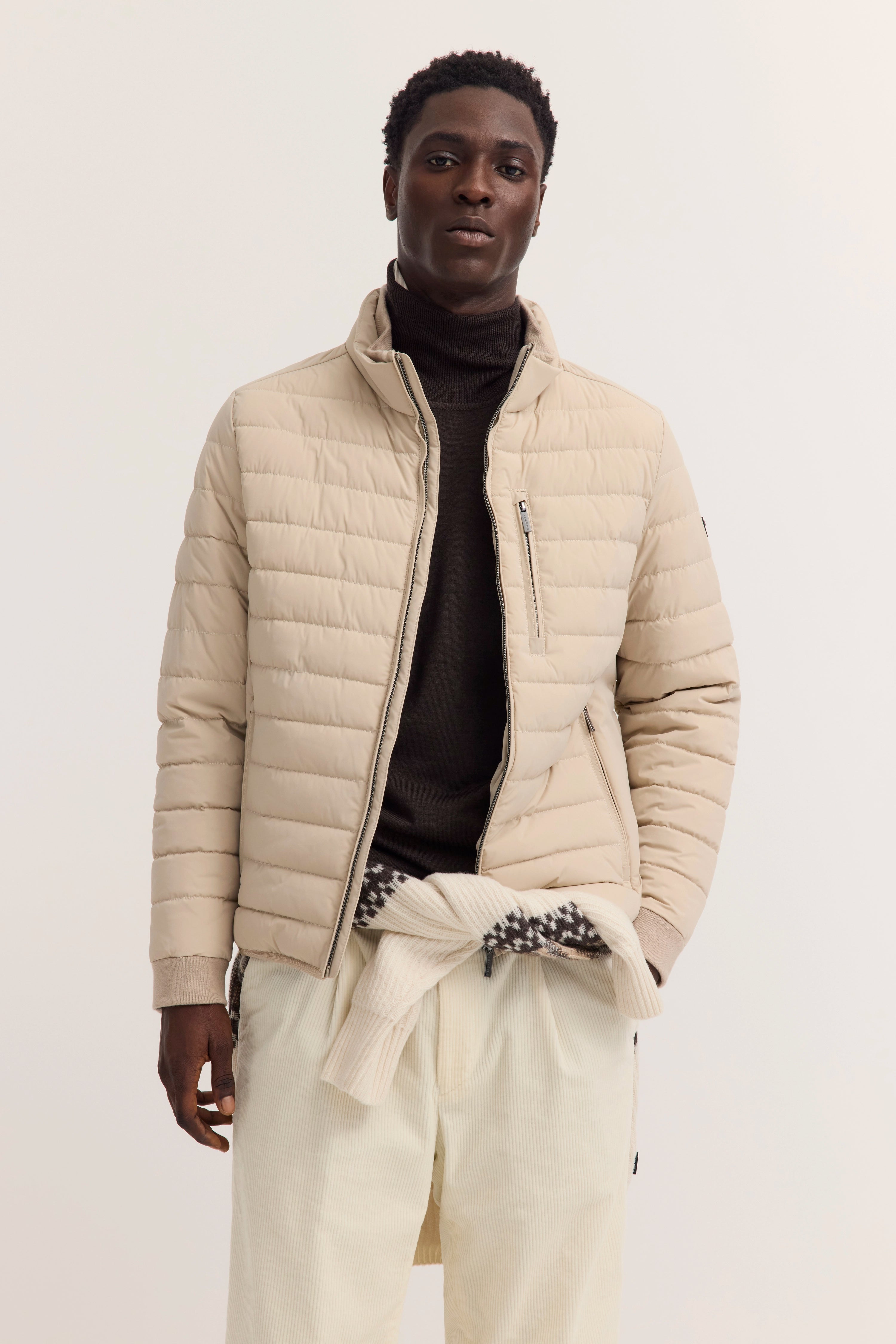 Beiges Pufferjacke-Outfit von bugatti, schwarzer Rollkragen, cremefarbene Hose, Model stehend, lässiges Posen.