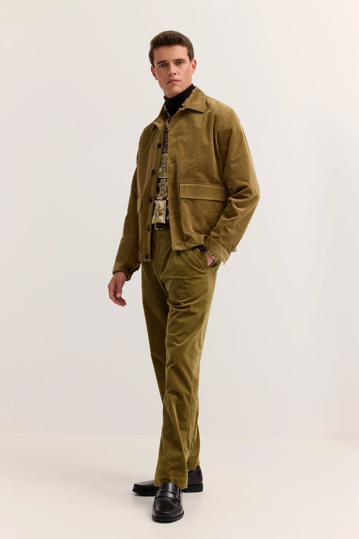 Model in olivgrünem Cord-Outfit mit gemustertem Hemd, schwarze Schuhe, entspannte Haltung.