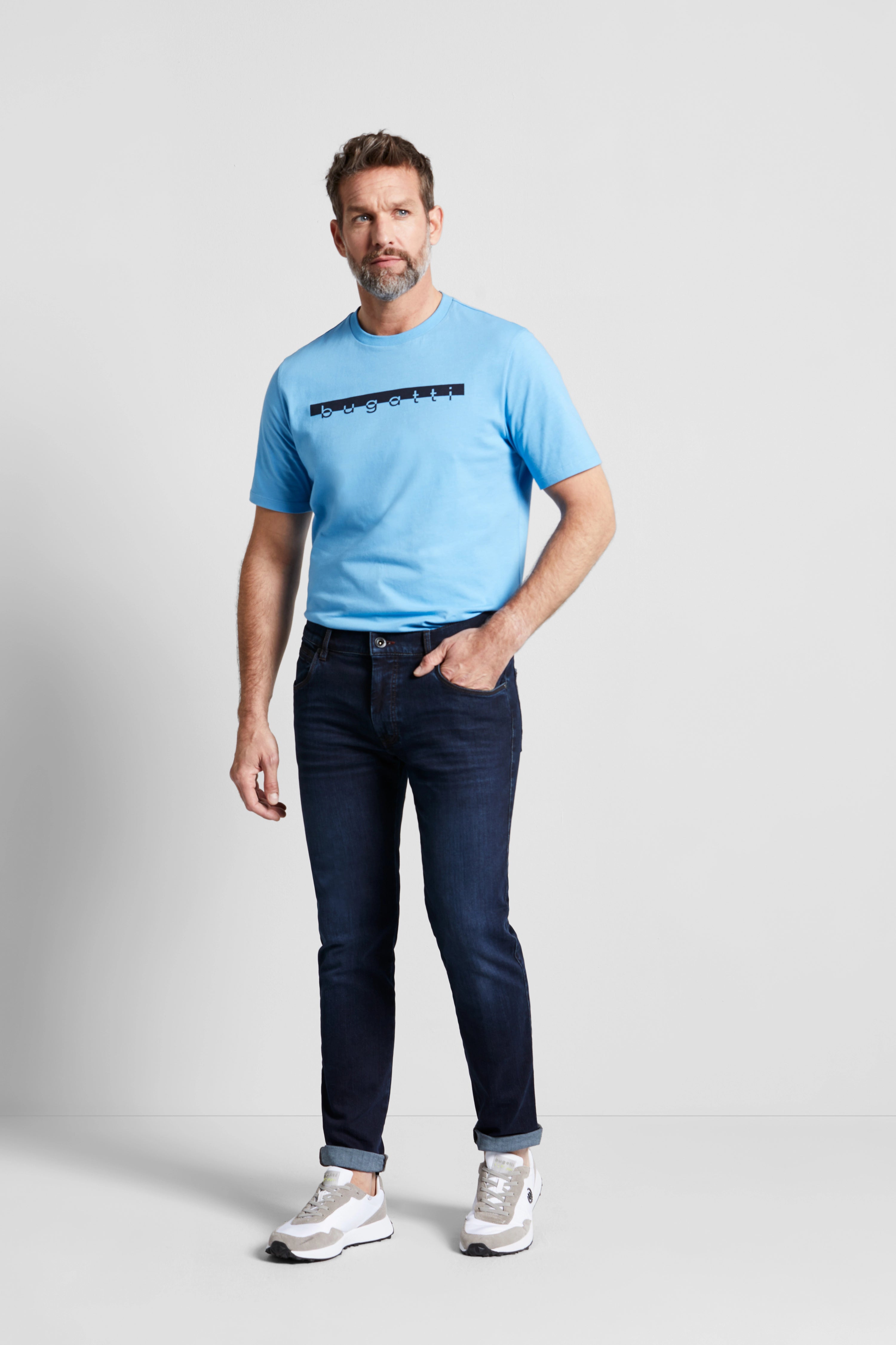 Model trägt bugatti blaues T-Shirt und dunkle Jeans. Lässig-stehende Pose, urbaner Stil.