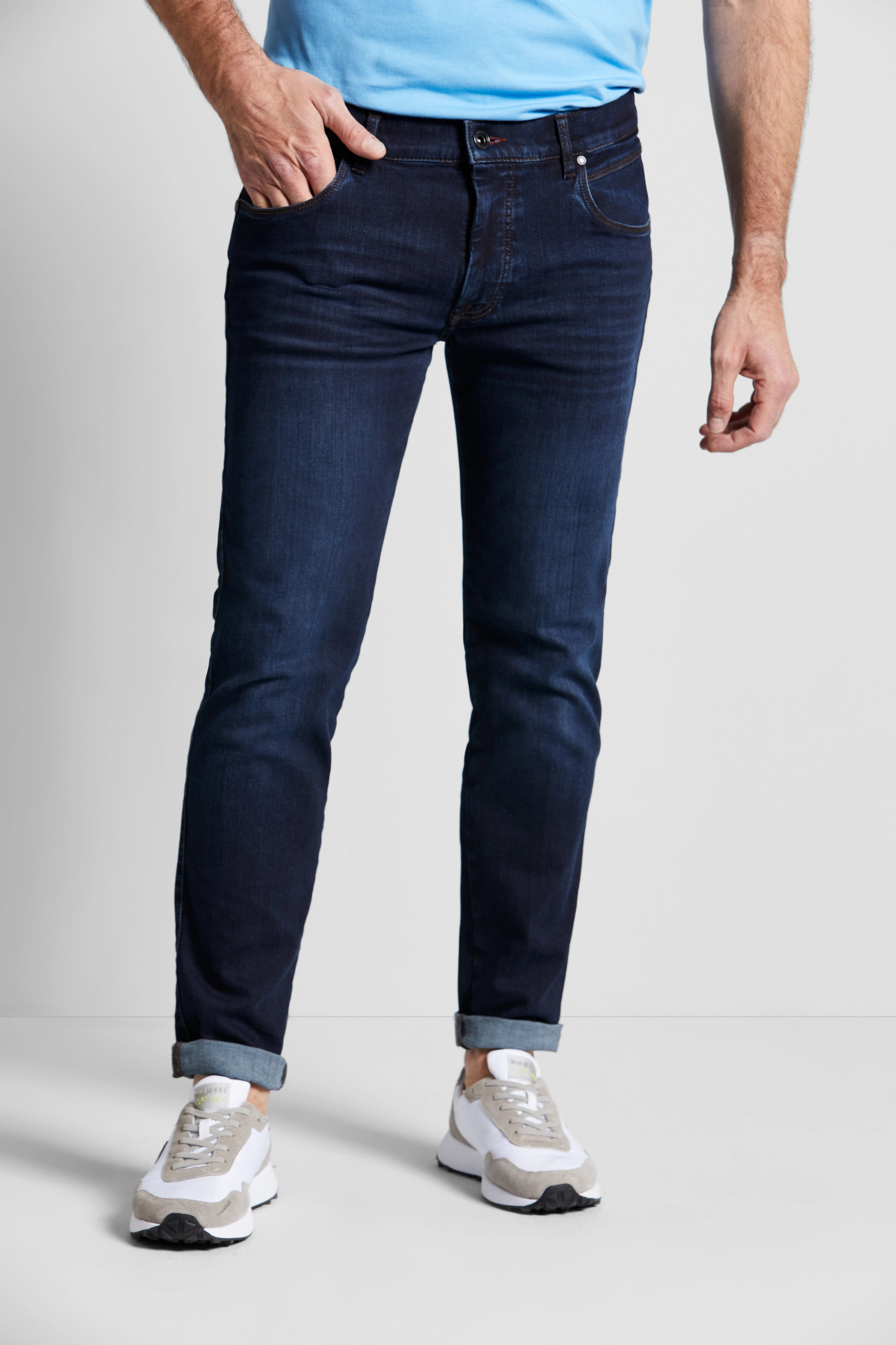 Dunkelblaue bugatti Jeans, leger getragen, mit hellblauem Shirt und weißen Sneakern. Modelbild, lässige Pose.