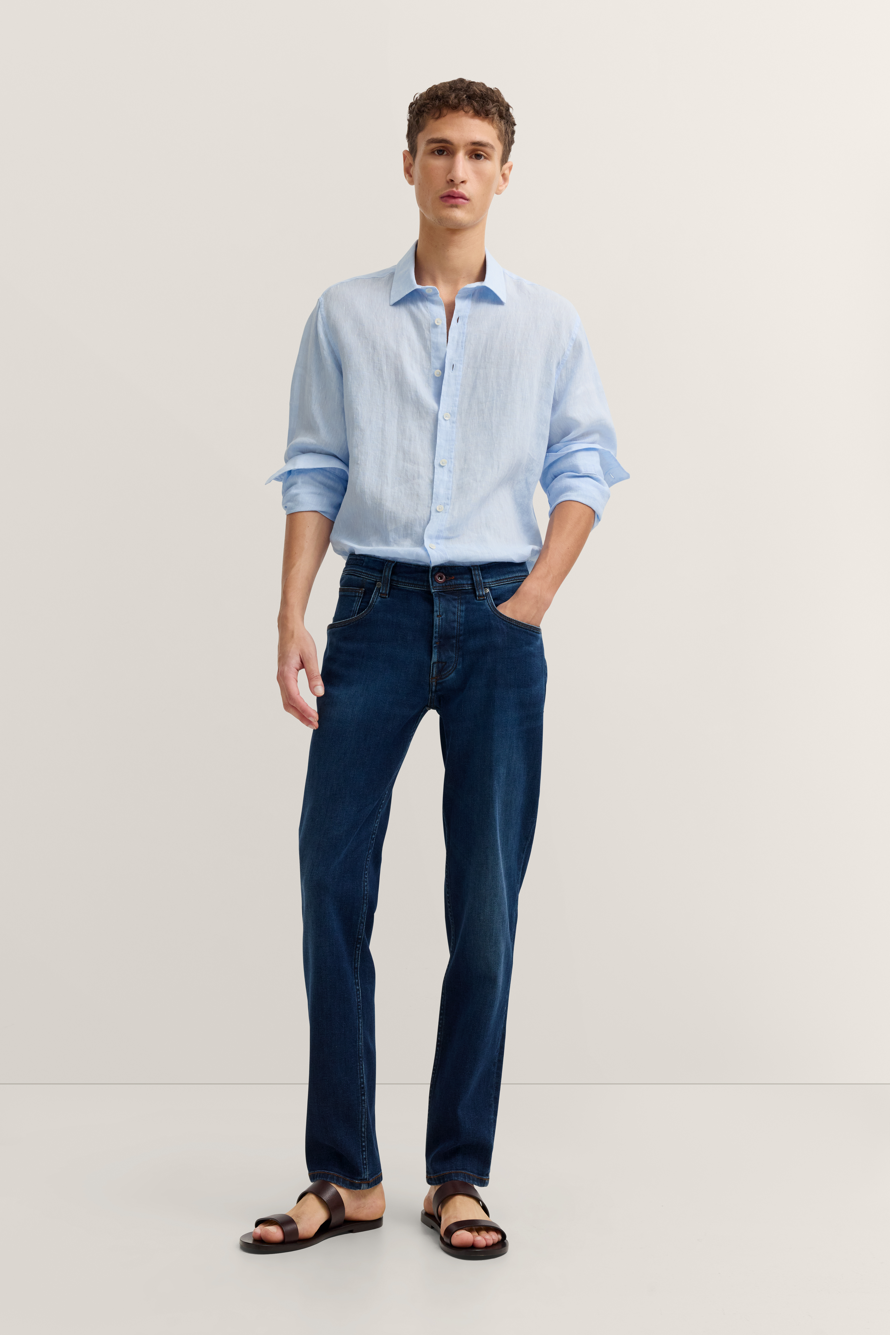 Model in hellblauem Hemd, dunkler Jeans und Sandalen. Steht entspannt, Hände in Hosentaschen.