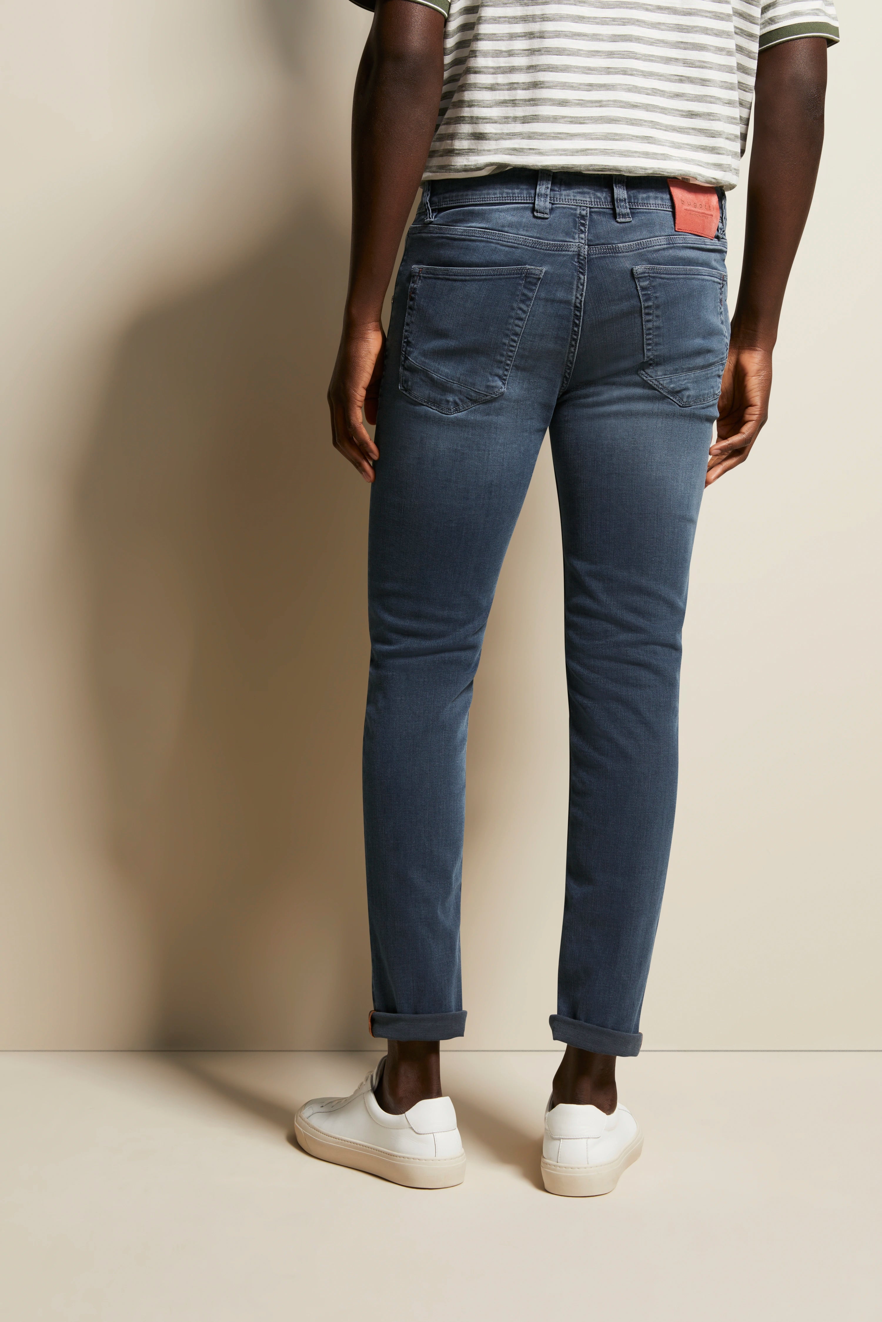 Jeans in Blau, Modelbild von hinten, lässig stehend, weißes Streifenshirt, weiße Sneaker.