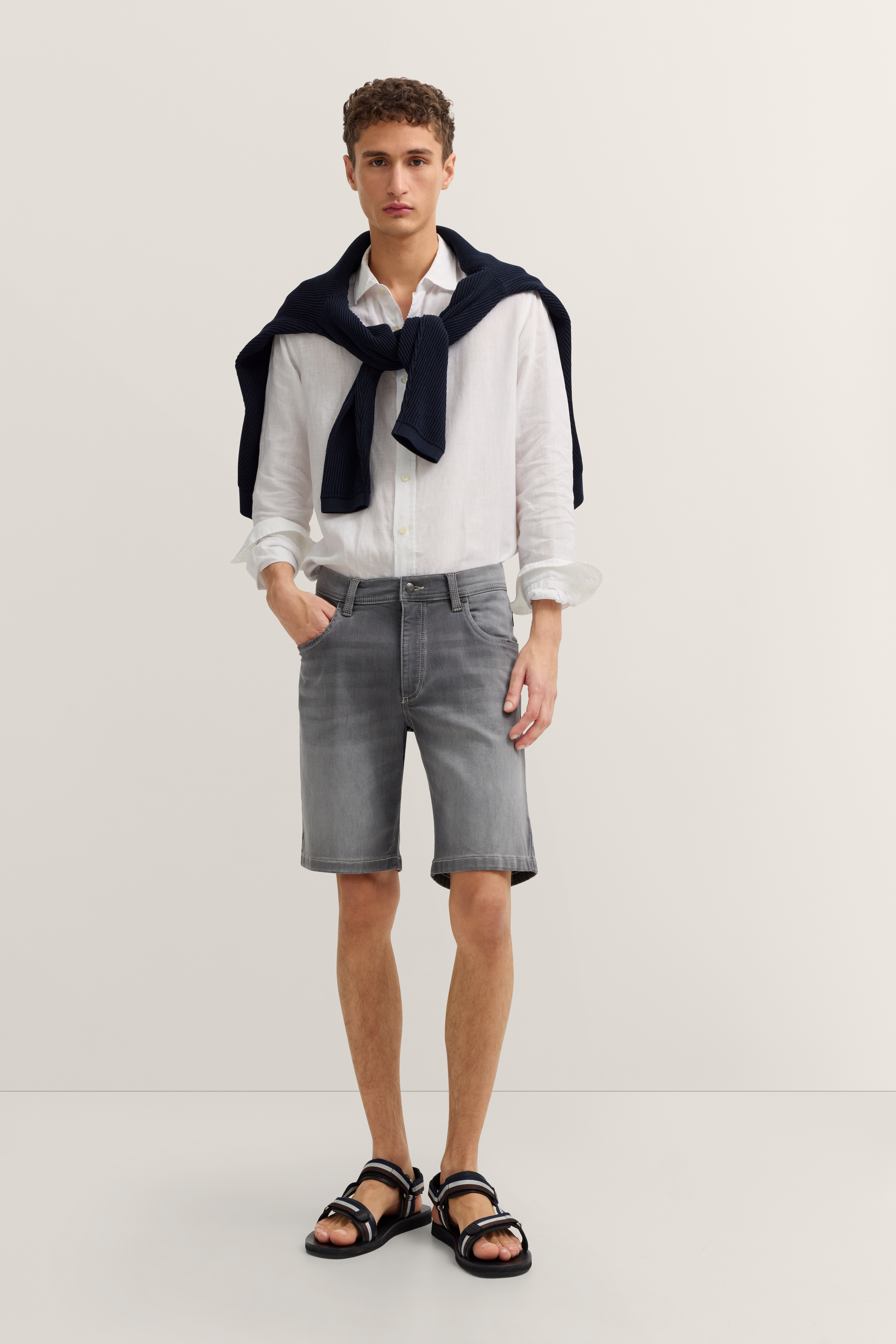 Model trägt weißes Hemd, graue Shorts, dunkelblauen Pullover und Sandalen. Lässige Pose. bugatti Stil.