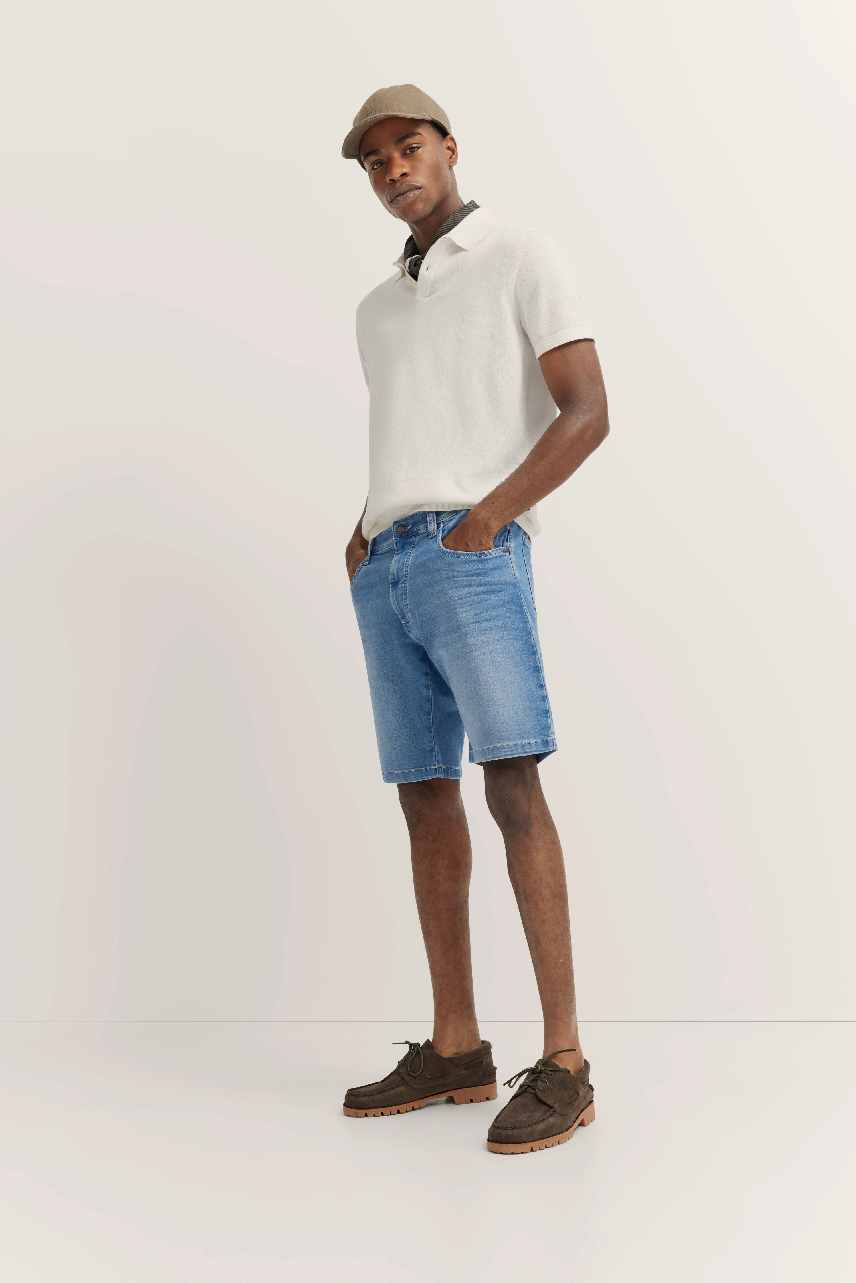 Modelbild: Weißes Poloshirt, blaue Jeansshorts, braune Schuhe, beiger Hut. Lässige Pose.