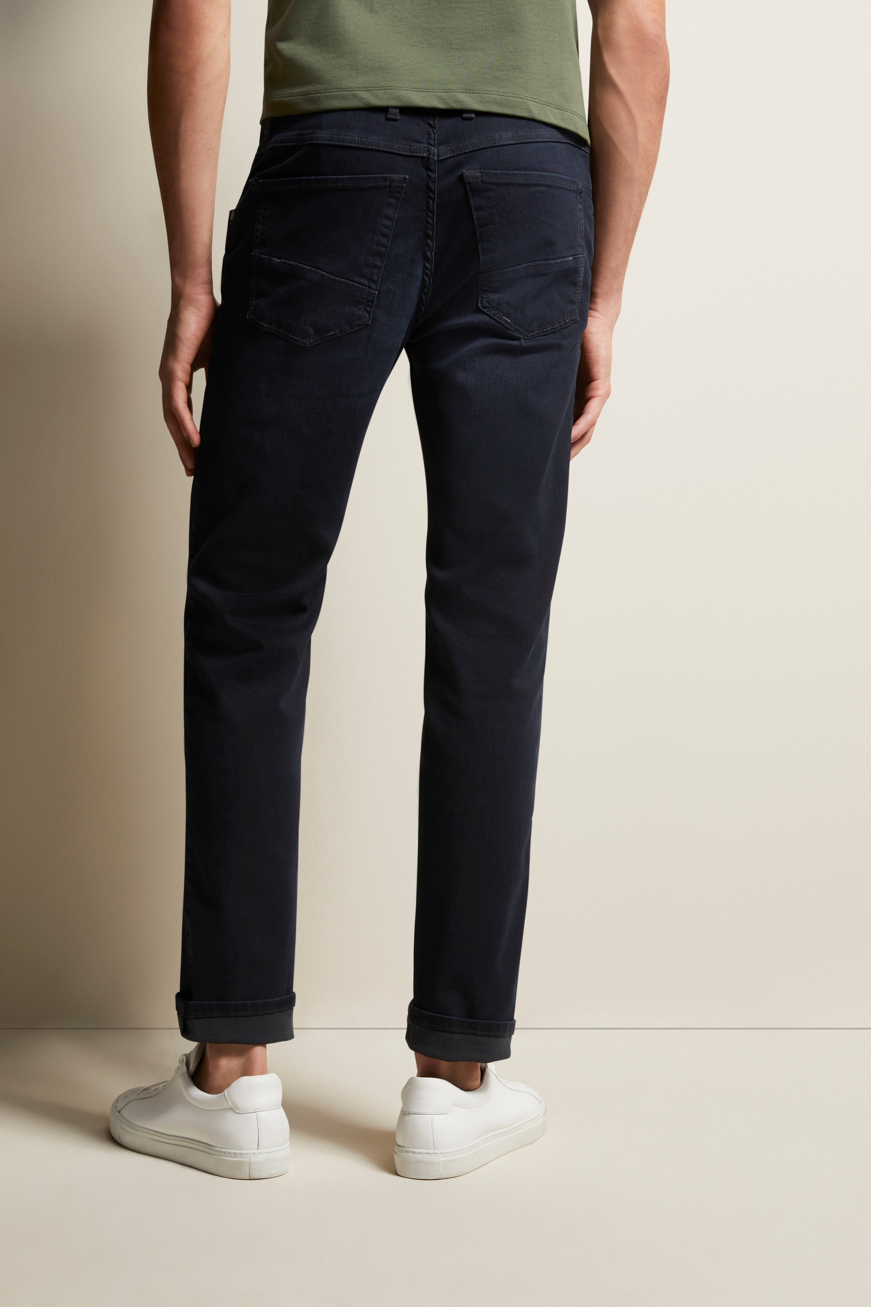 Donkerblauwe jeans, casual, met witte sneaker en olijfgroen shirt. Model van achteren, ontspannen staand.