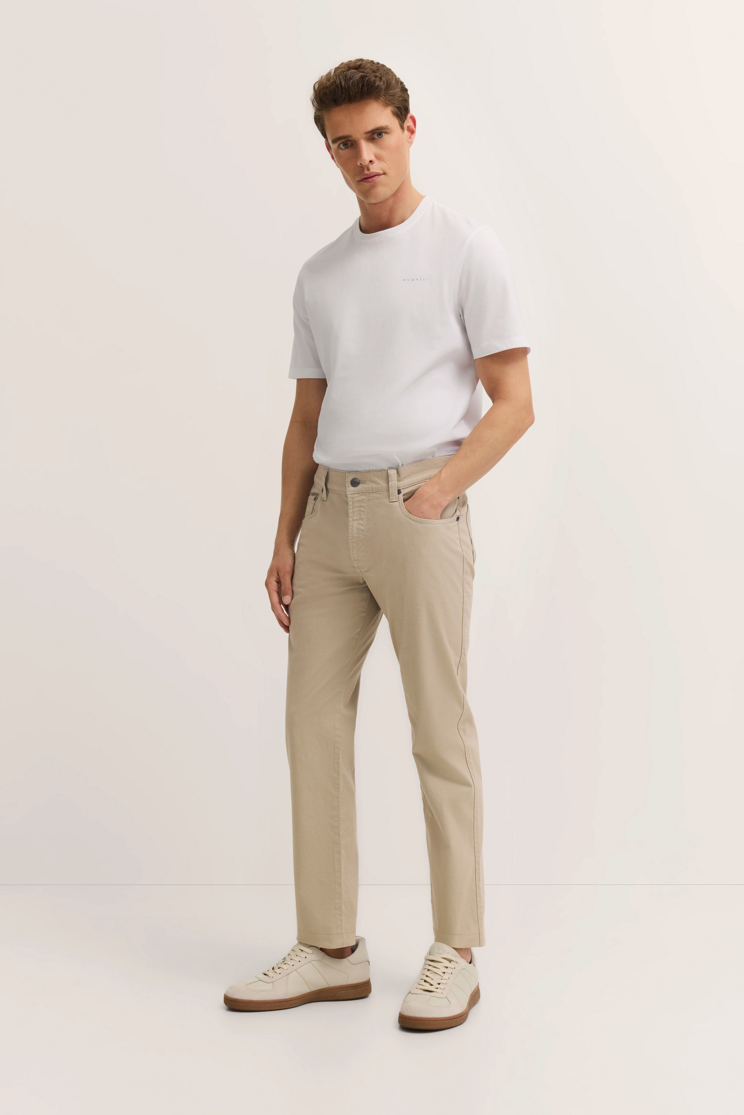Model in wit bugatti T-shirt, beige broek en sneakers, ontspannen houding.