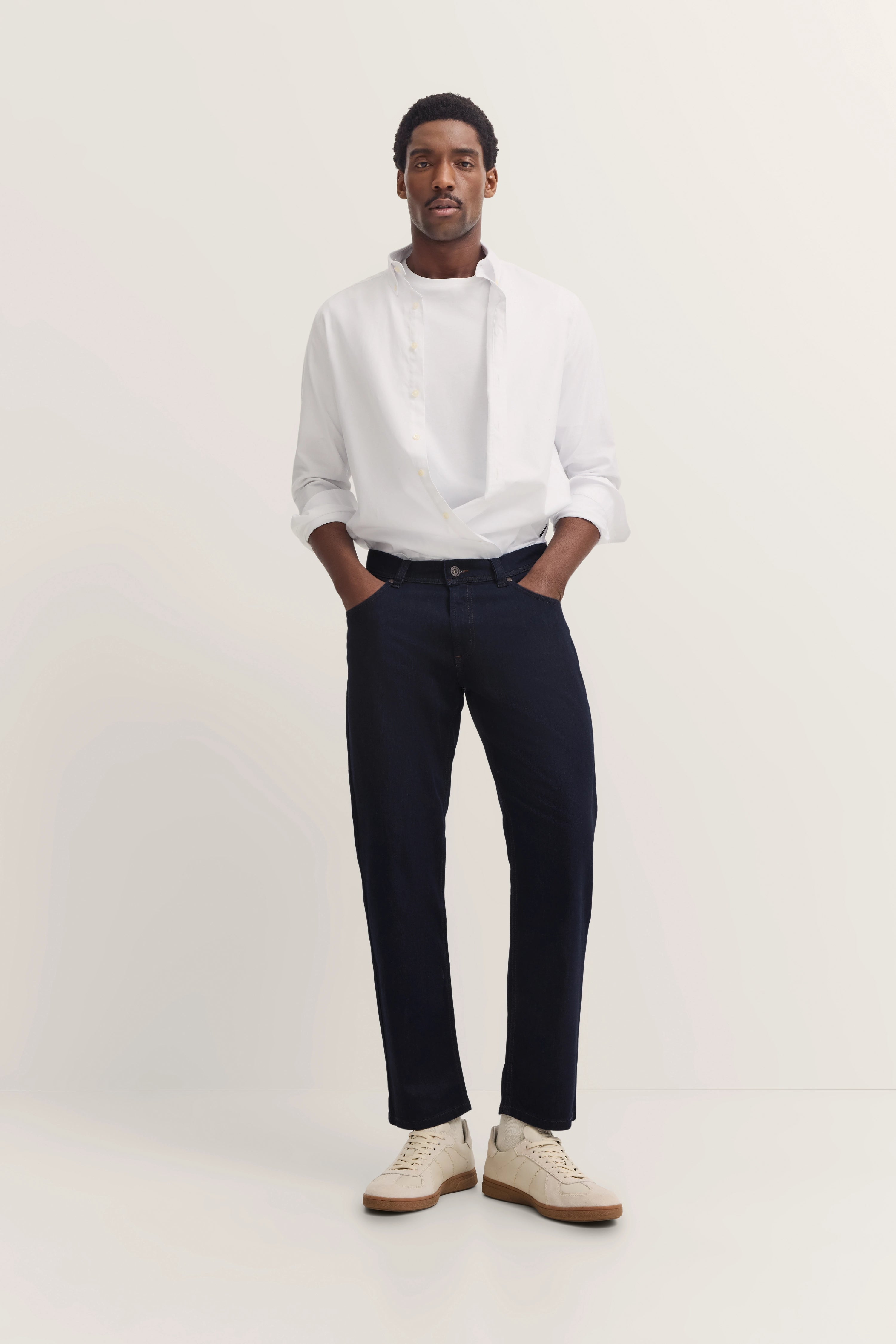Model trägt weißes Hemd, dunkle Jeans und beige Sneaker von bugatti, lässige Pose, Hände in den Taschen.