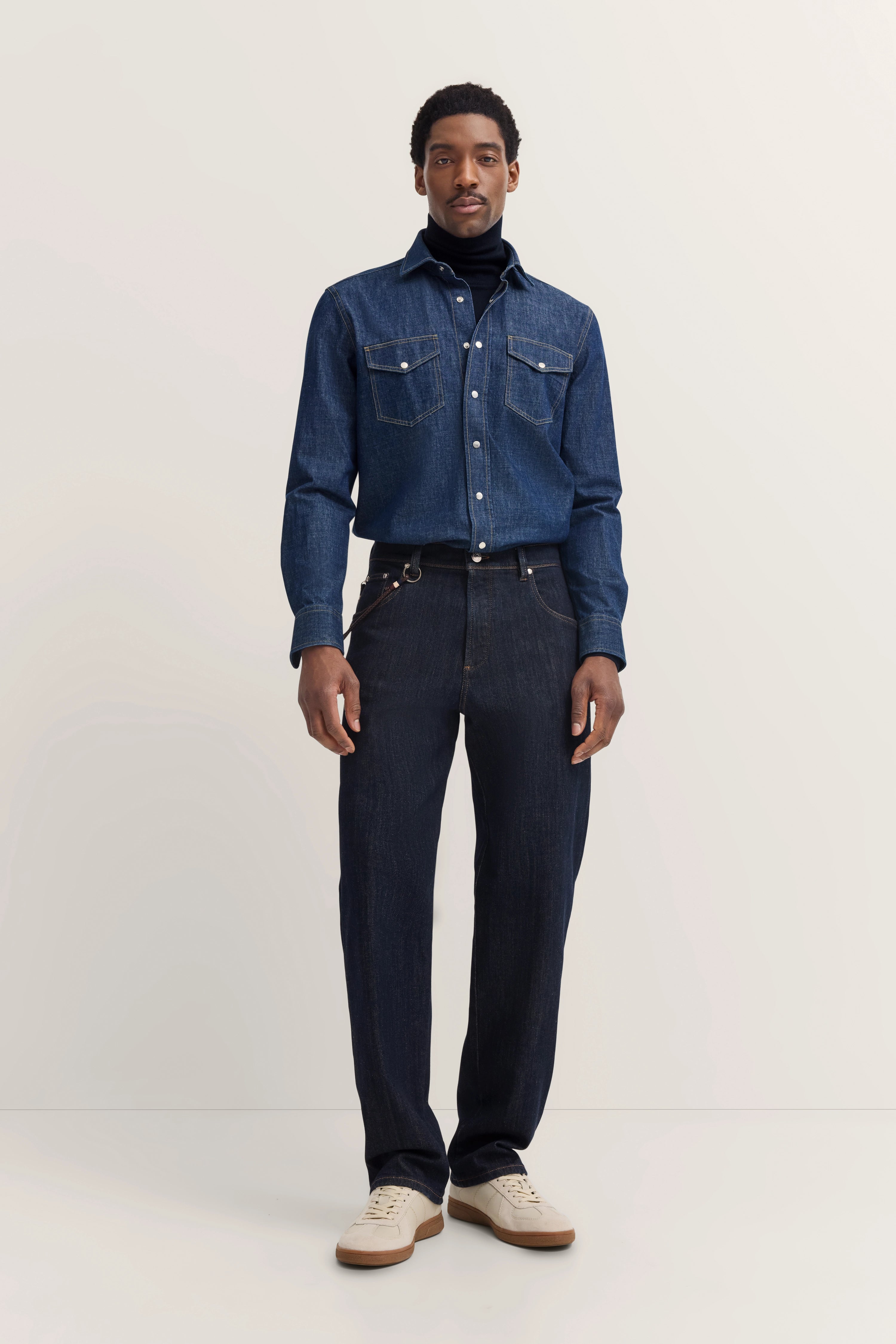 Model in Denimhemd und Jeans von bugatti, blau, mit schwarzem Rollkragenpullover, lässige Pose.