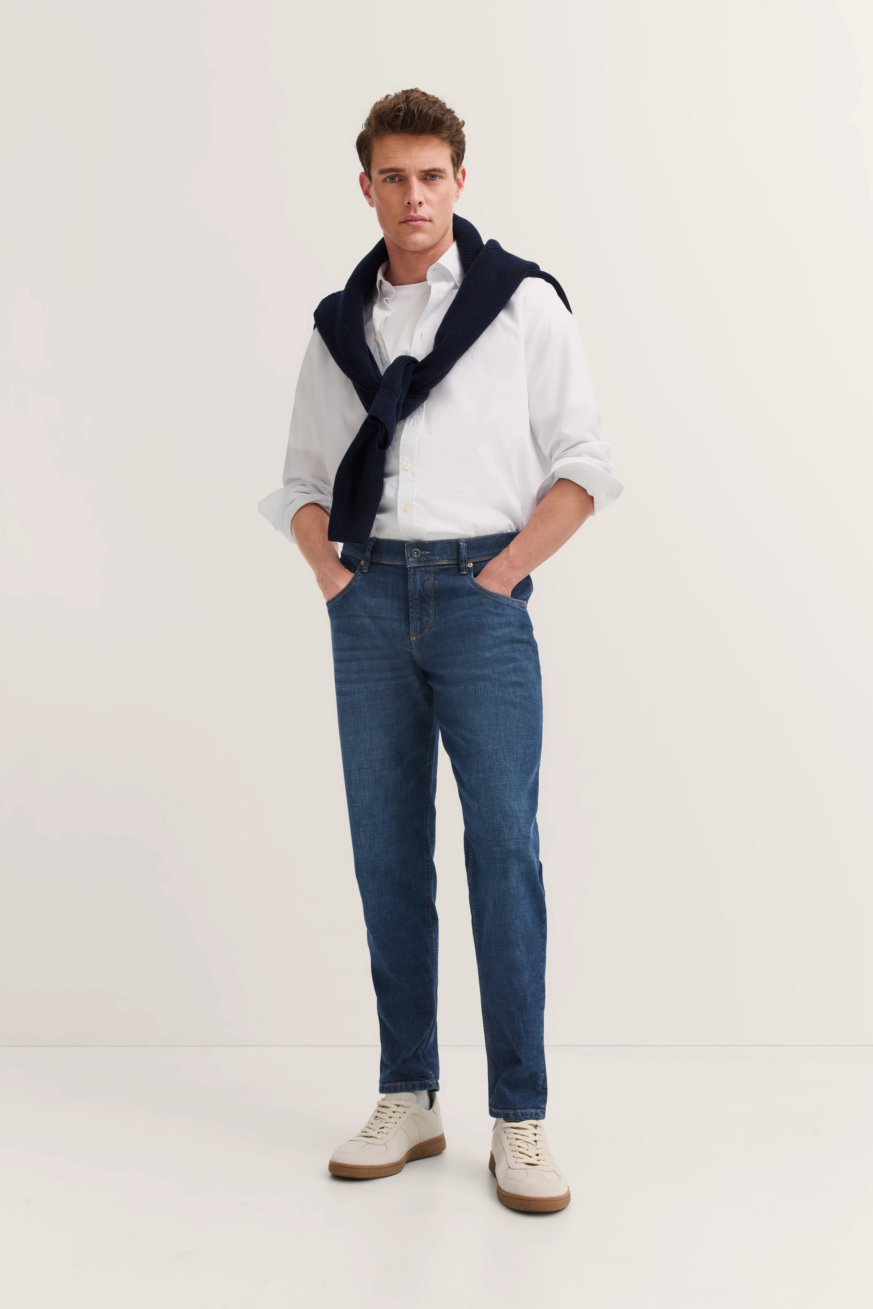 Weißes Hemd, blaue Jeans, dunkelblauer Pullover über Schultern, Model stehend, Hände in Taschen.