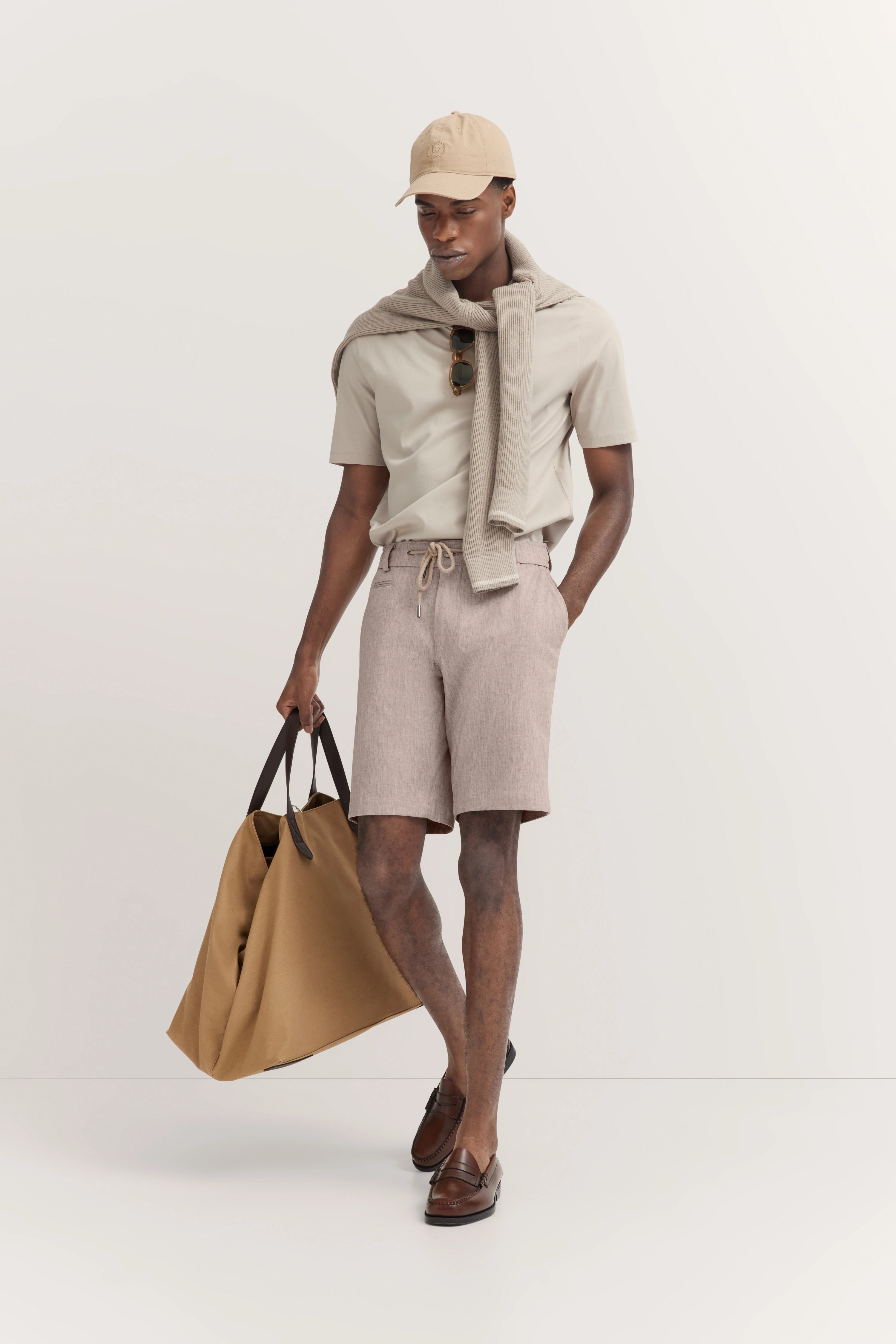 Model in Beige-T-Shirt und Shorts, Pullover über Schultern, beige Cap, Tasche. Lässig, bugatti Stil.