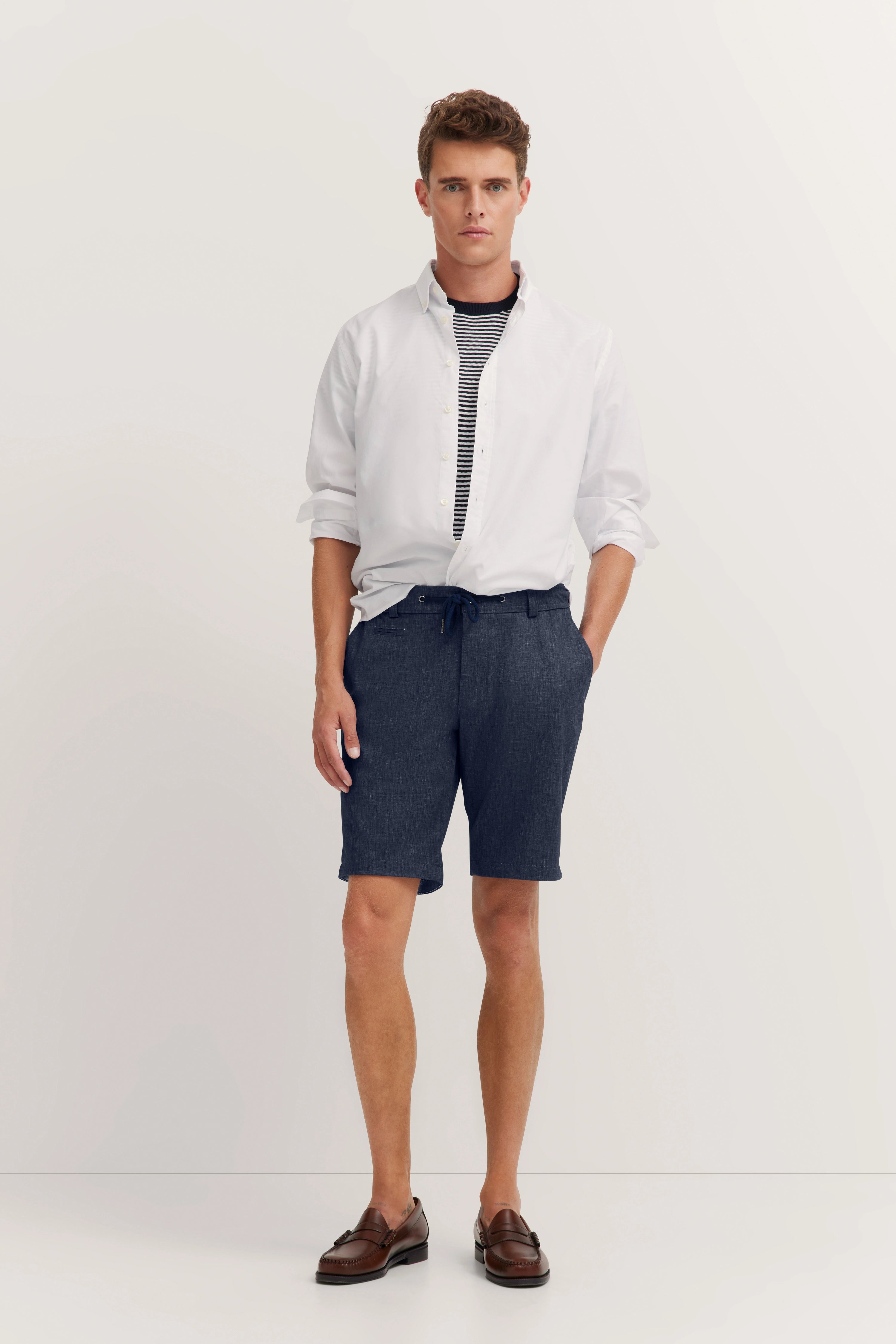 Model in weißem Hemd, gestreiftem Shirt, dunkelblauen Shorts, braunen Schuhen, lässiger Stand.