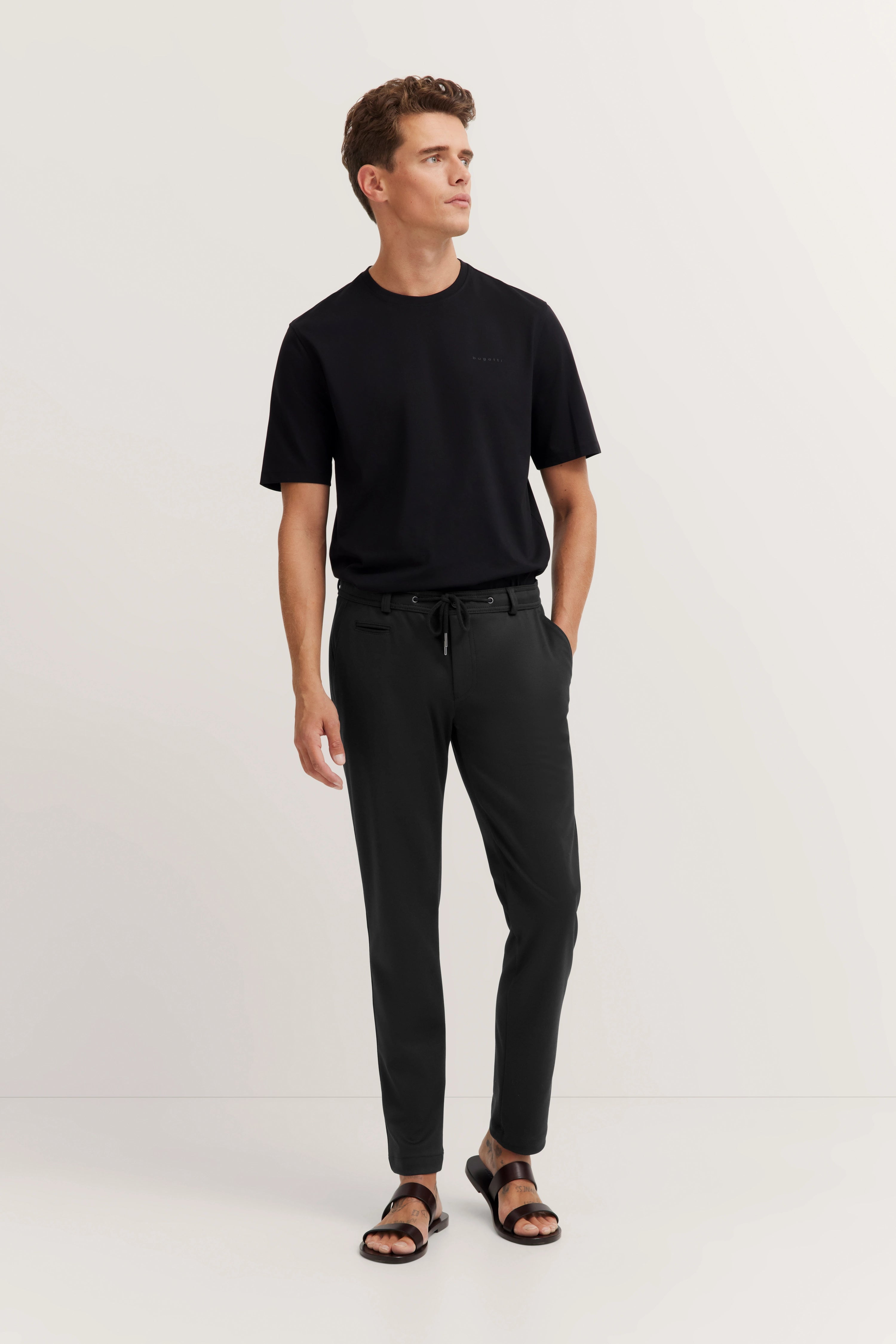 Model in schwarzem bugatti T-Shirt und Hose, legeres Posing, Blick zur Seite.