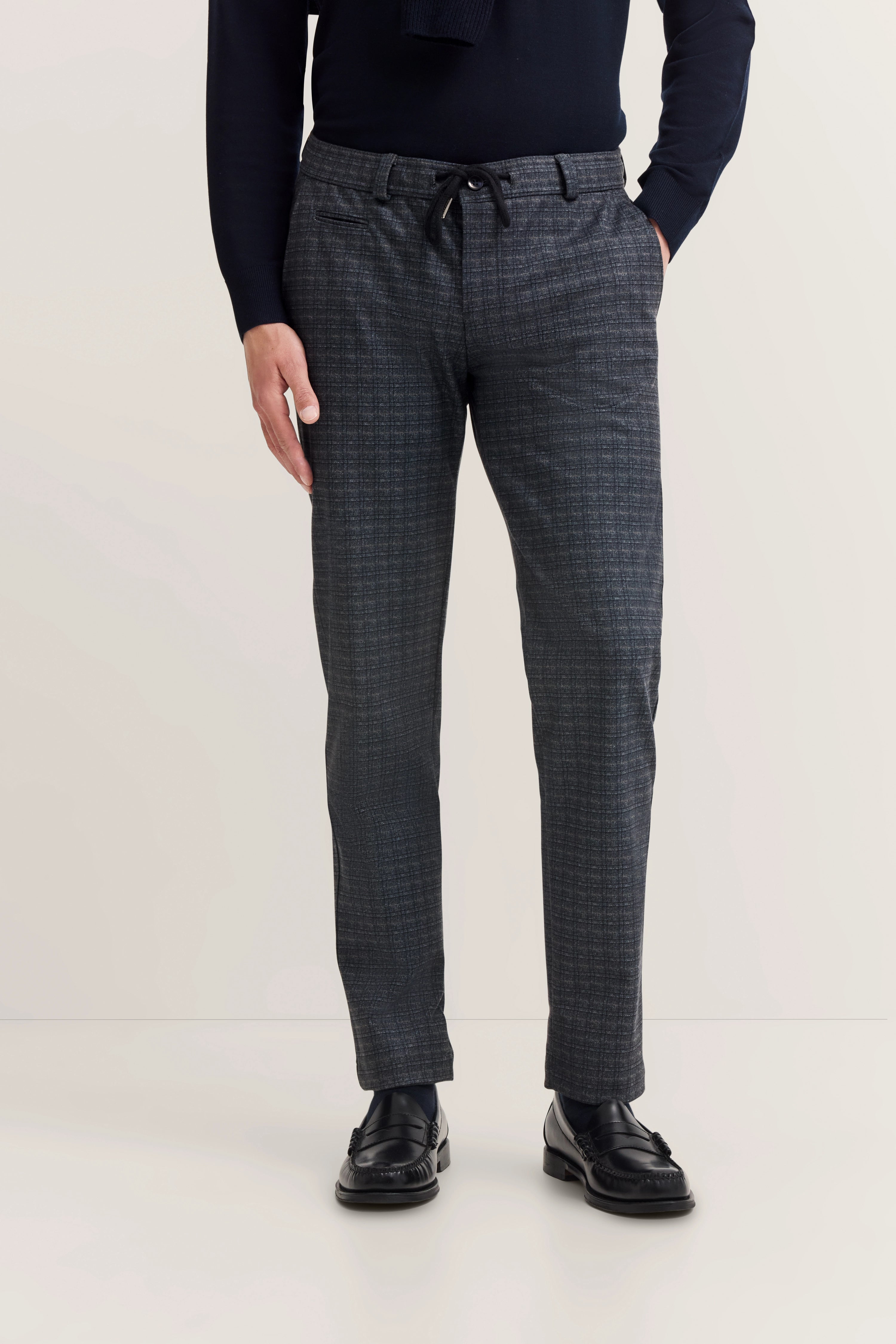 Graue karierte Hose, legeres Modelbild, Hände in Taschen, schwarzer Oberteil und Schuhe von bugatti.