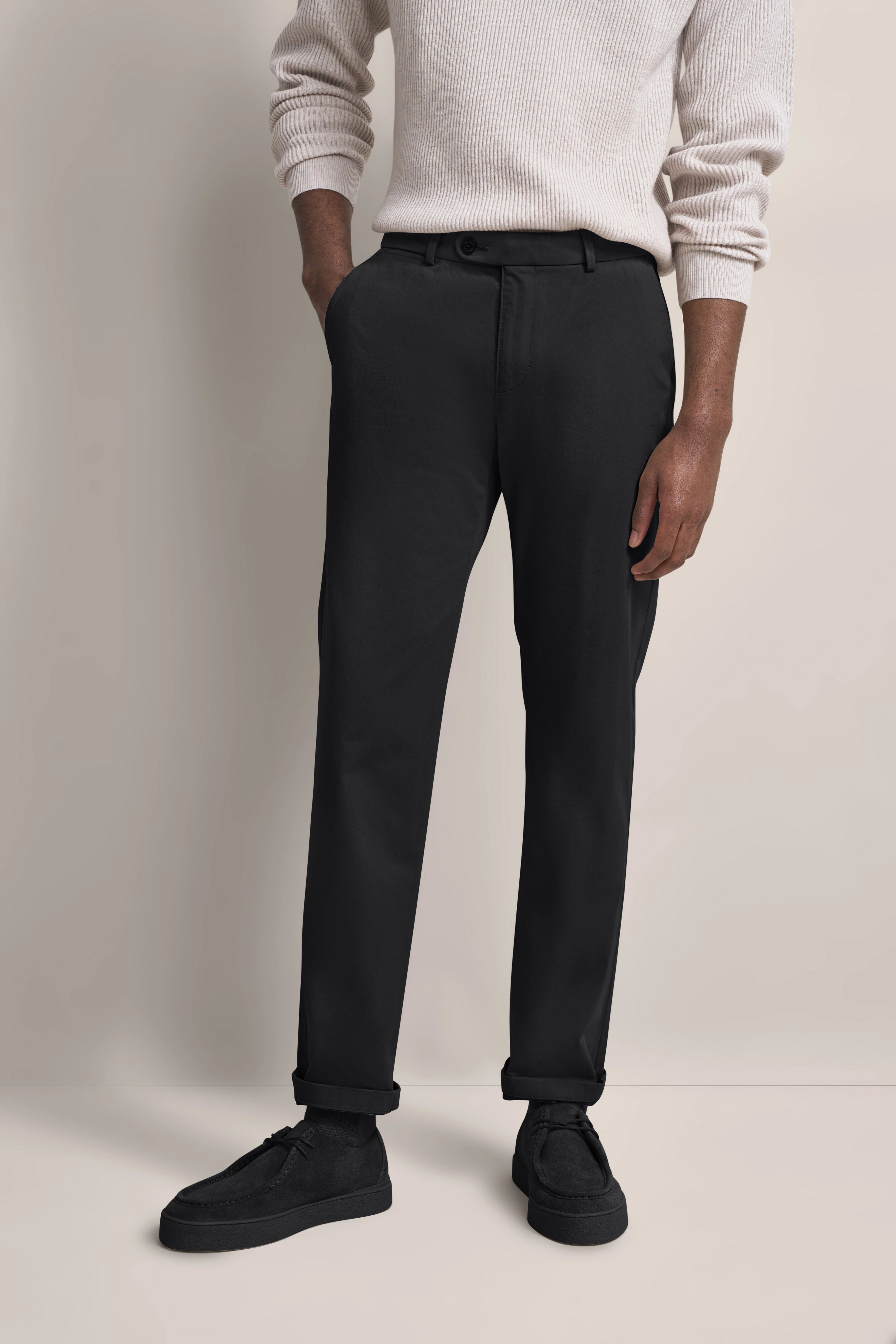 bugatti Modelbild: Weißer Pullover, schwarze Hose, schwarze Schuhe, legeres Posing, Hände in Taschen.
