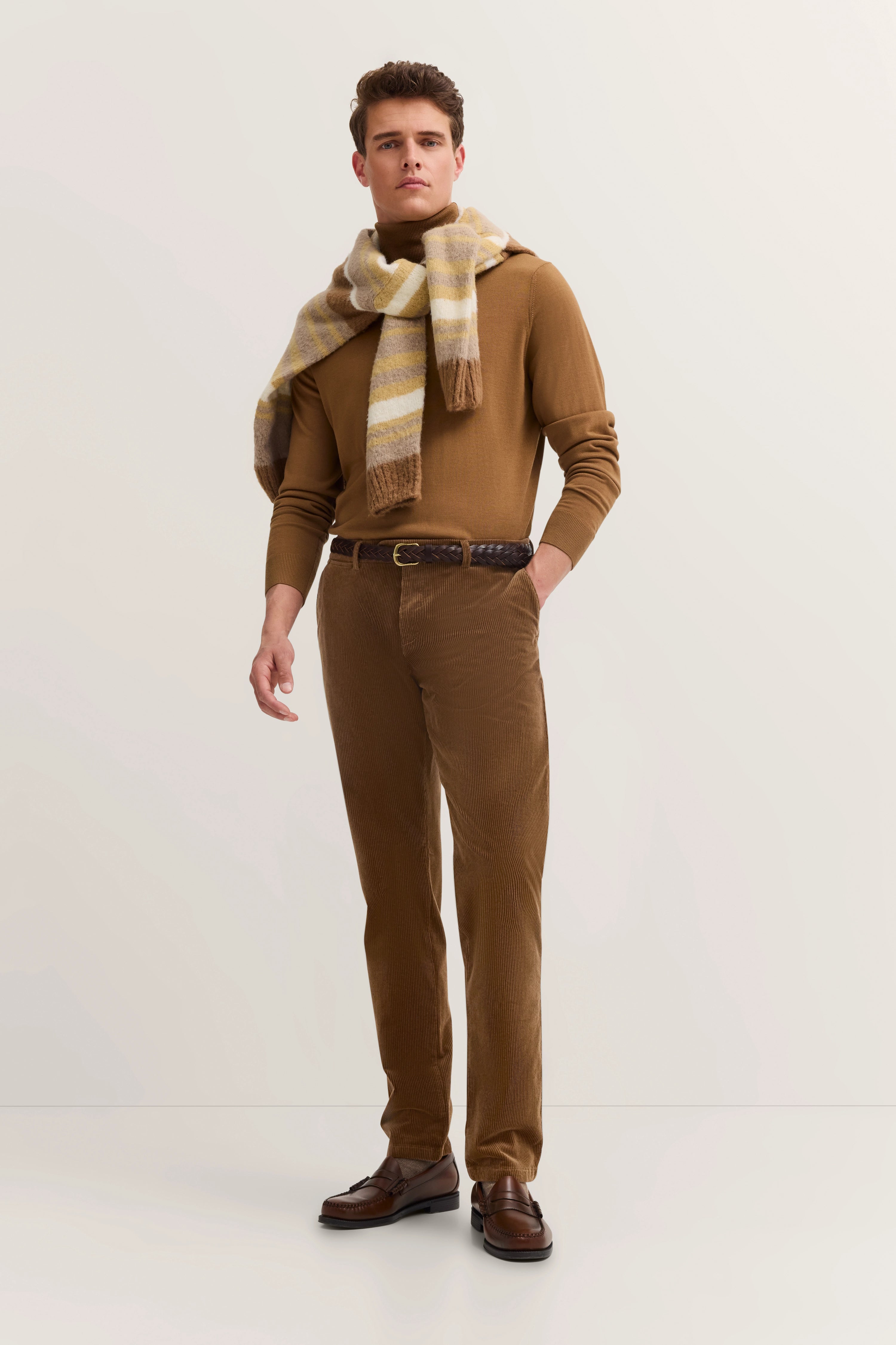 Männermodell in braunem Rollkragenpullover, Cordhose, gestreiftem Schal, lässige Pose.